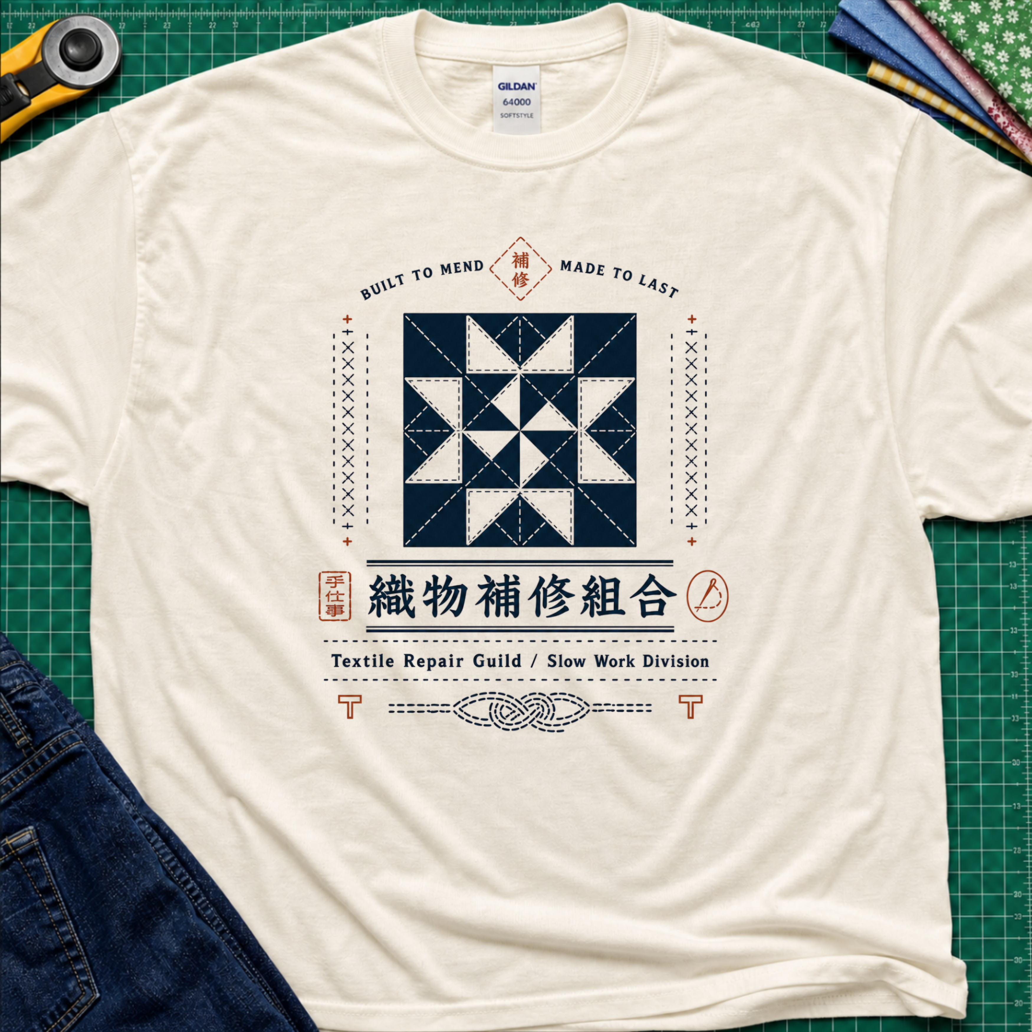 A light cream Gildan t-shirt with a central graphic of a quilt block and Japanese text reading 'Textile Repair Guild / Slow Work Division'. The shirt is laid flat on a green cutting mat with a rotary cutter, fabric stack and blue jeans partially visible in the corners.