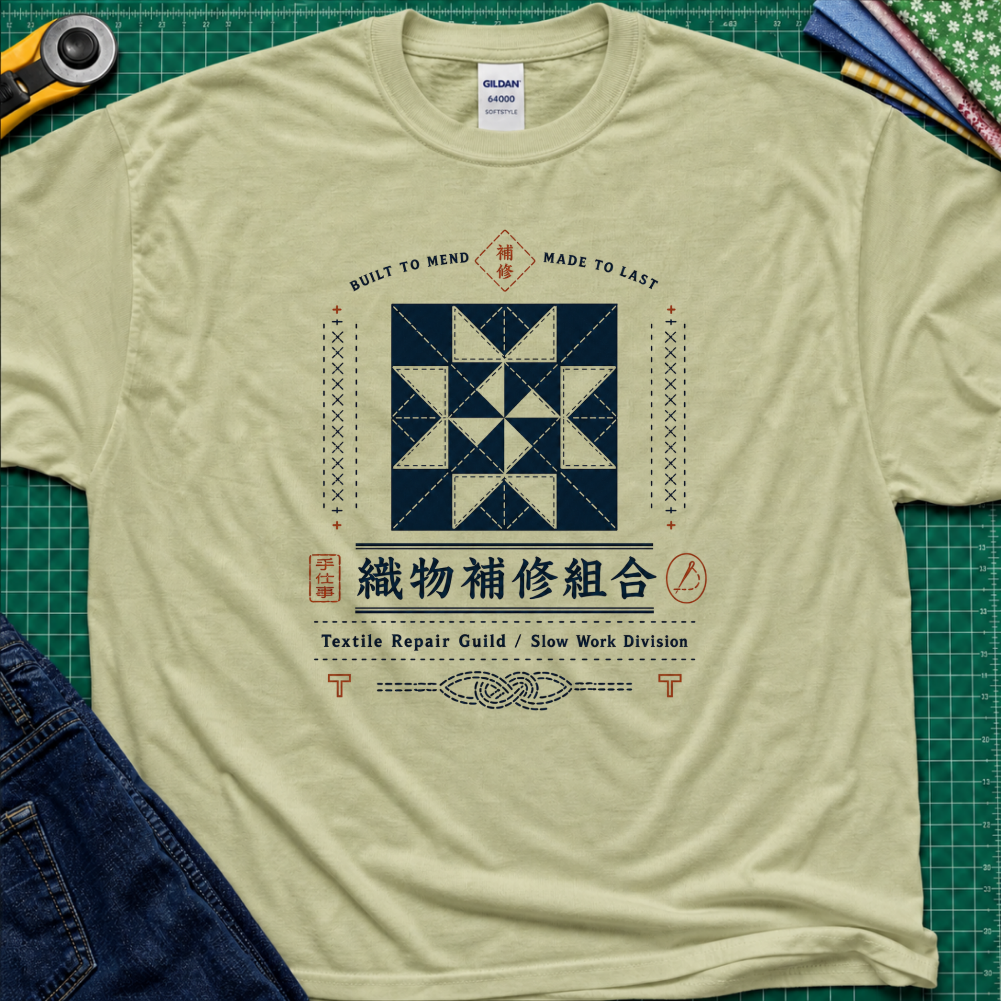 A light pistachio green Gildan t-shirt with a central graphic of a quilt block and Japanese text reading 'Textile Repair Guild / Slow Work Division'. The shirt is laid flat on a green cutting mat with a rotary cutter, fabric stack and blue jeans partially visible in the corners.