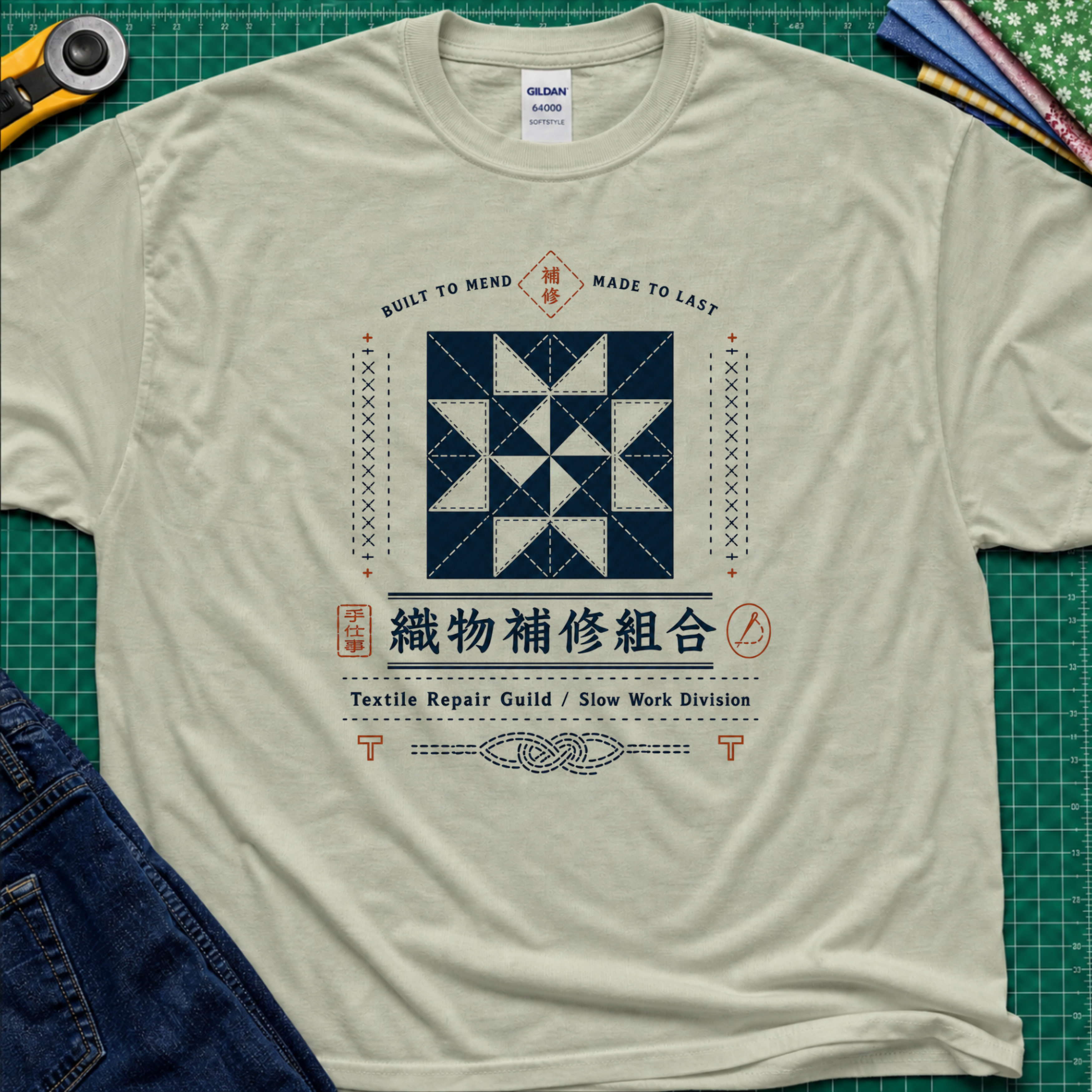 A light sage green Gildan t-shirt with a central graphic of a quilt block and Japanese text reading 'Textile Repair Guild / Slow Work Division'. The shirt is laid flat on a green cutting mat with a rotary cutter, fabric stack and blue jeans partially visible in the corners.