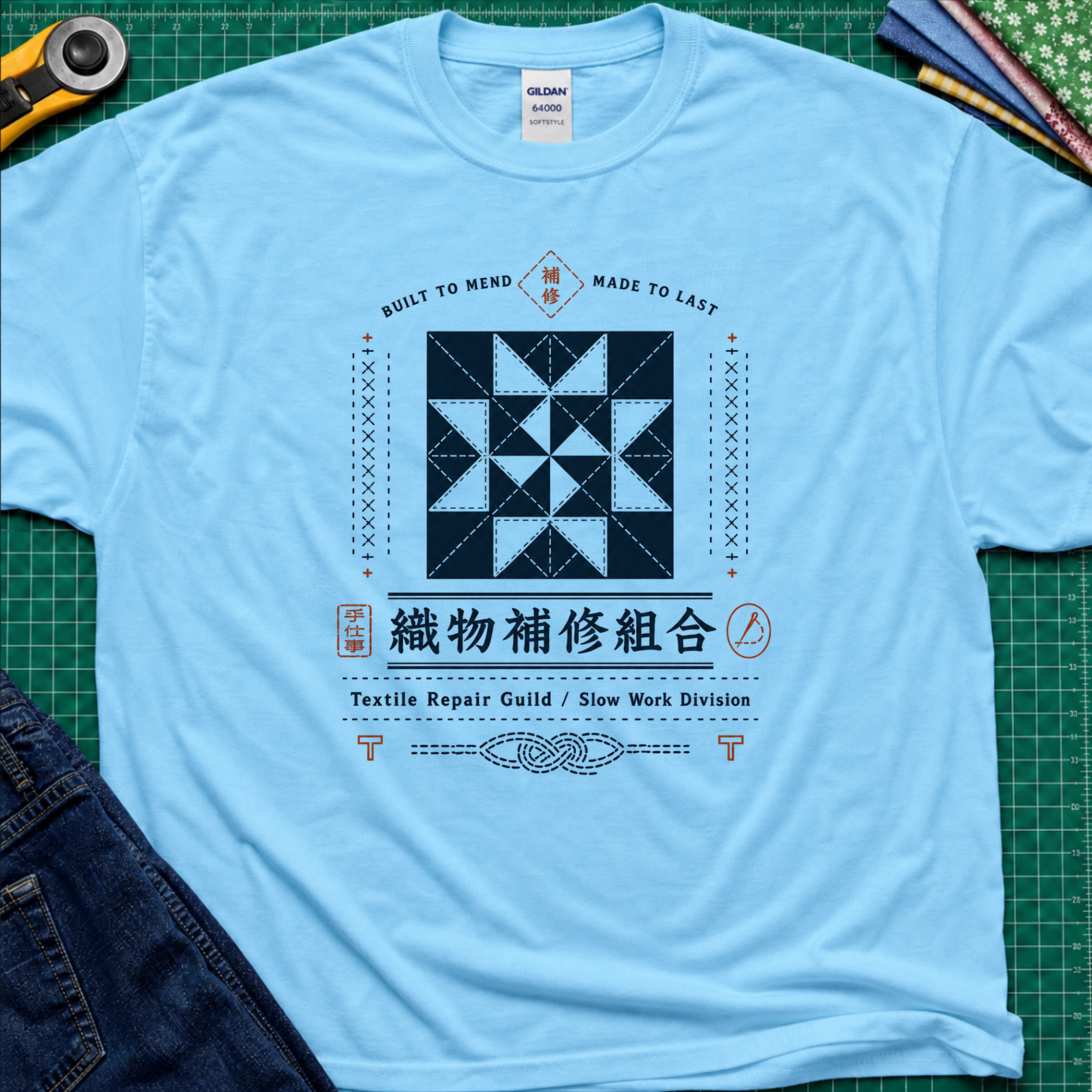 A sky blue Gildan t-shirt with a central graphic of a quilt block and Japanese text reading 'Textile Repair Guild / Slow Work Division'. The shirt is laid flat on a green cutting mat with a rotary cutter, fabric stack and blue jeans partially visible in the corners.