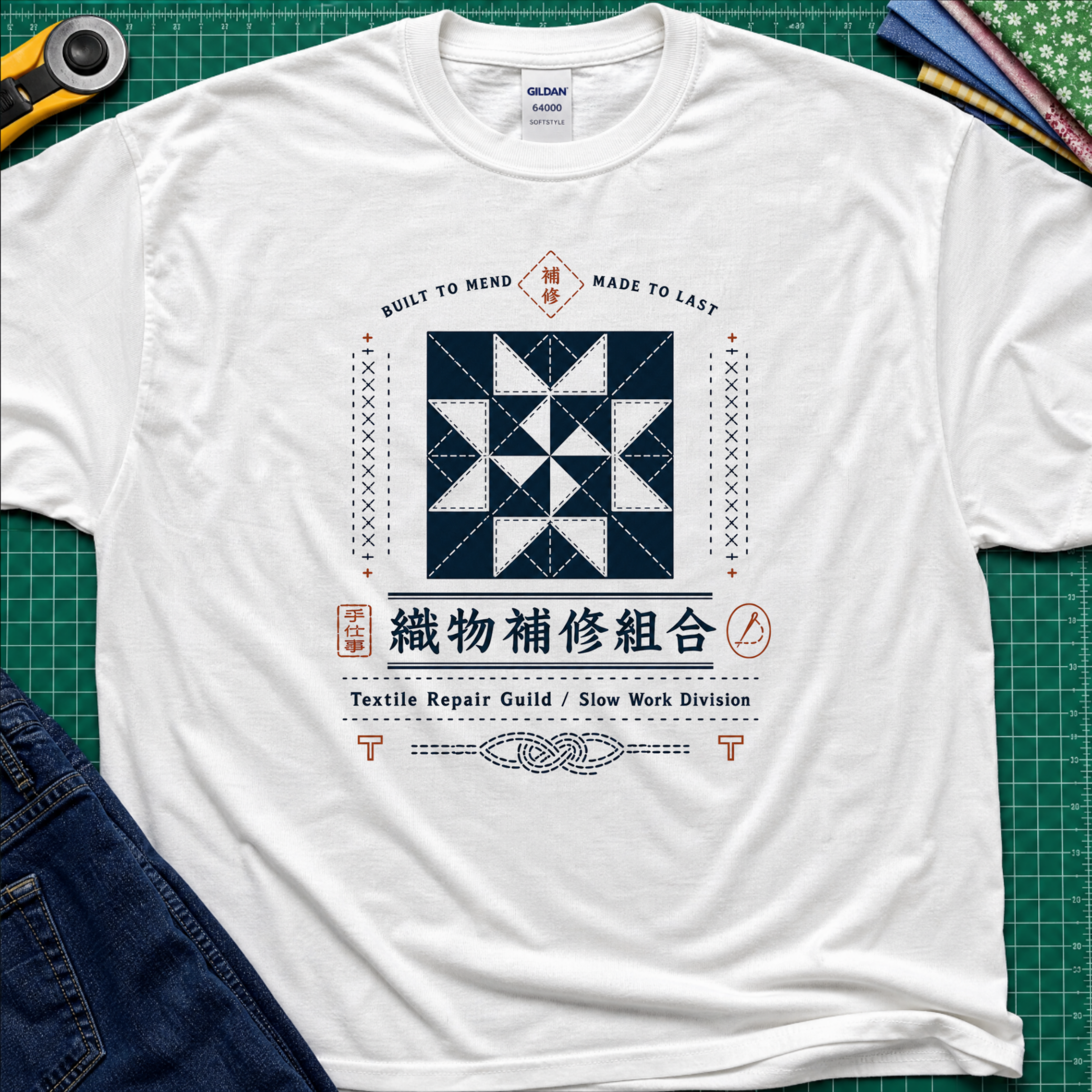 A white Gildan t-shirt with a central graphic of a quilt block and Japanese text reading 'Textile Repair Guild / Slow Work Division'. The shirt is laid flat on a green cutting mat with a rotary cutter, fabric stack and blue jeans partially visible in the corners.
