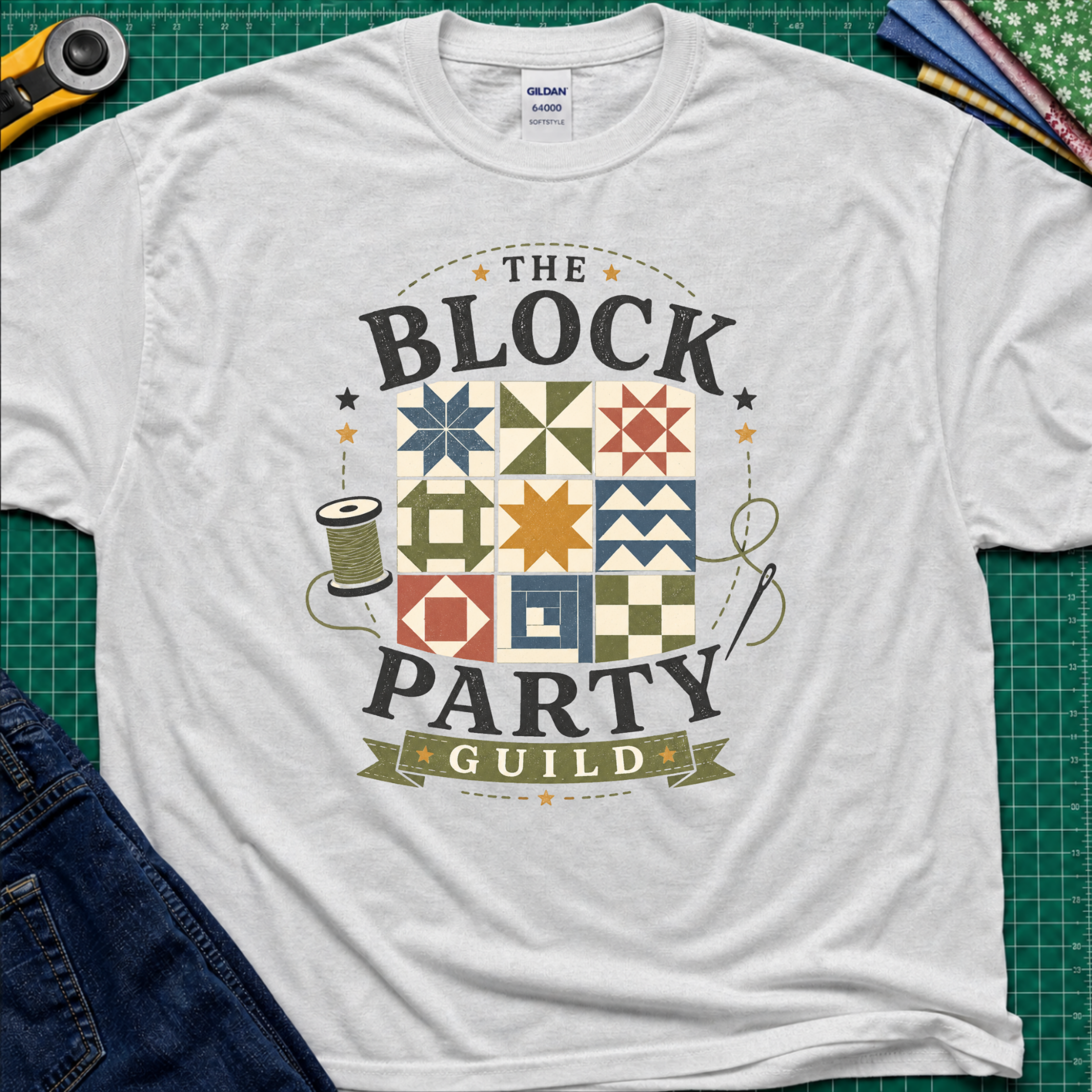 A light grey Gildan t-shirt with a vintage-style graphic reading 'THE BLOCK PARTY GUILD' around a set of six quilt blocks. The shirt is laid flat on a green cutting mat with sewing-themed props like a rotary cutter, fabric, and jeans partially visible in the corners.