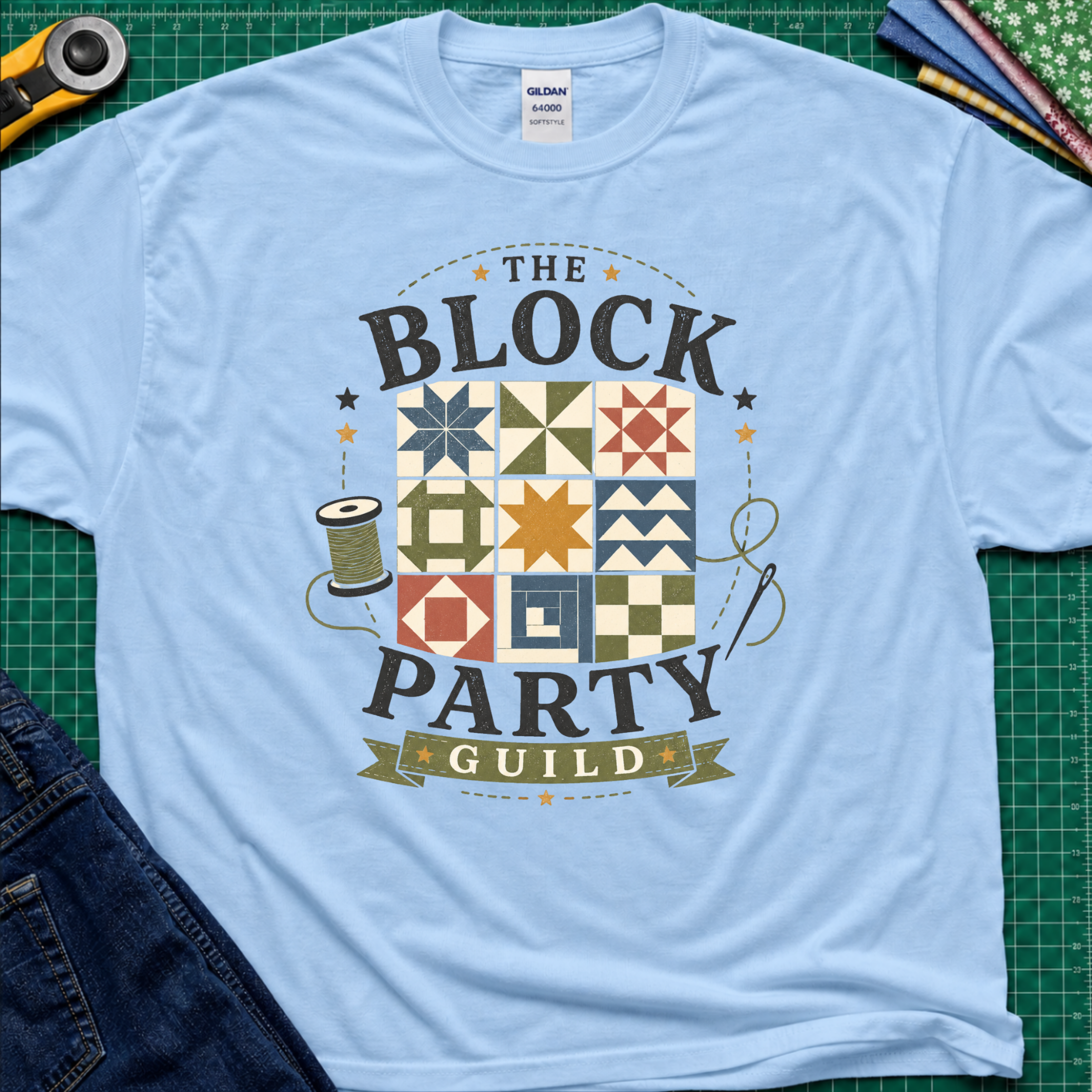 A light blue Gildan t-shirt with a vintage-style graphic reading 'THE BLOCK PARTY GUILD' around a set of six quilt blocks. The shirt is laid flat on a green cutting mat with sewing-themed props like a rotary cutter, fabric, and jeans partially visible in the corners.