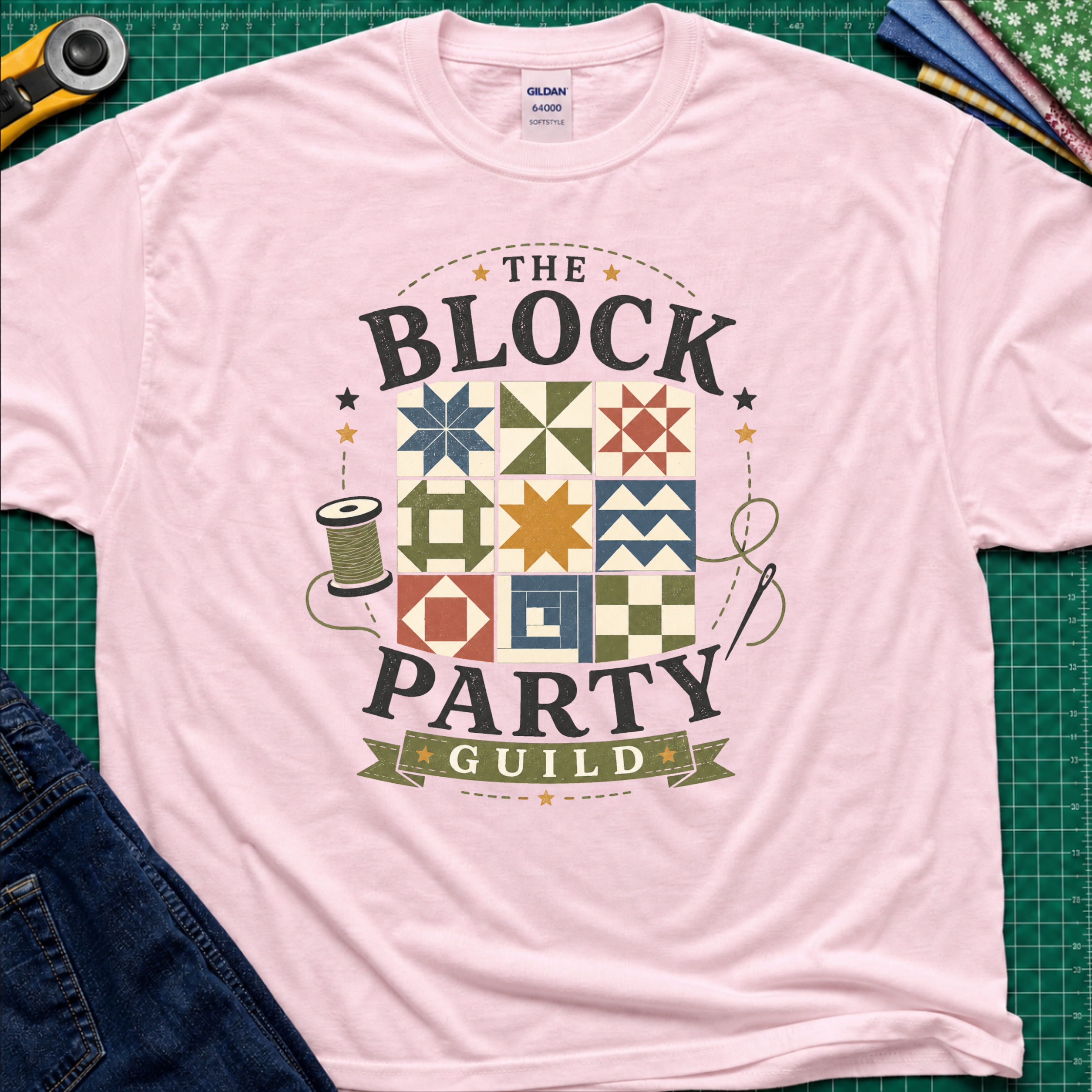 A light pink Gildan t-shirt with a vintage-style graphic reading 'THE BLOCK PARTY GUILD' around a set of six quilt blocks. The shirt is laid flat on a green cutting mat with sewing-themed props like a rotary cutter, fabric, and jeans partially visible in the corners.