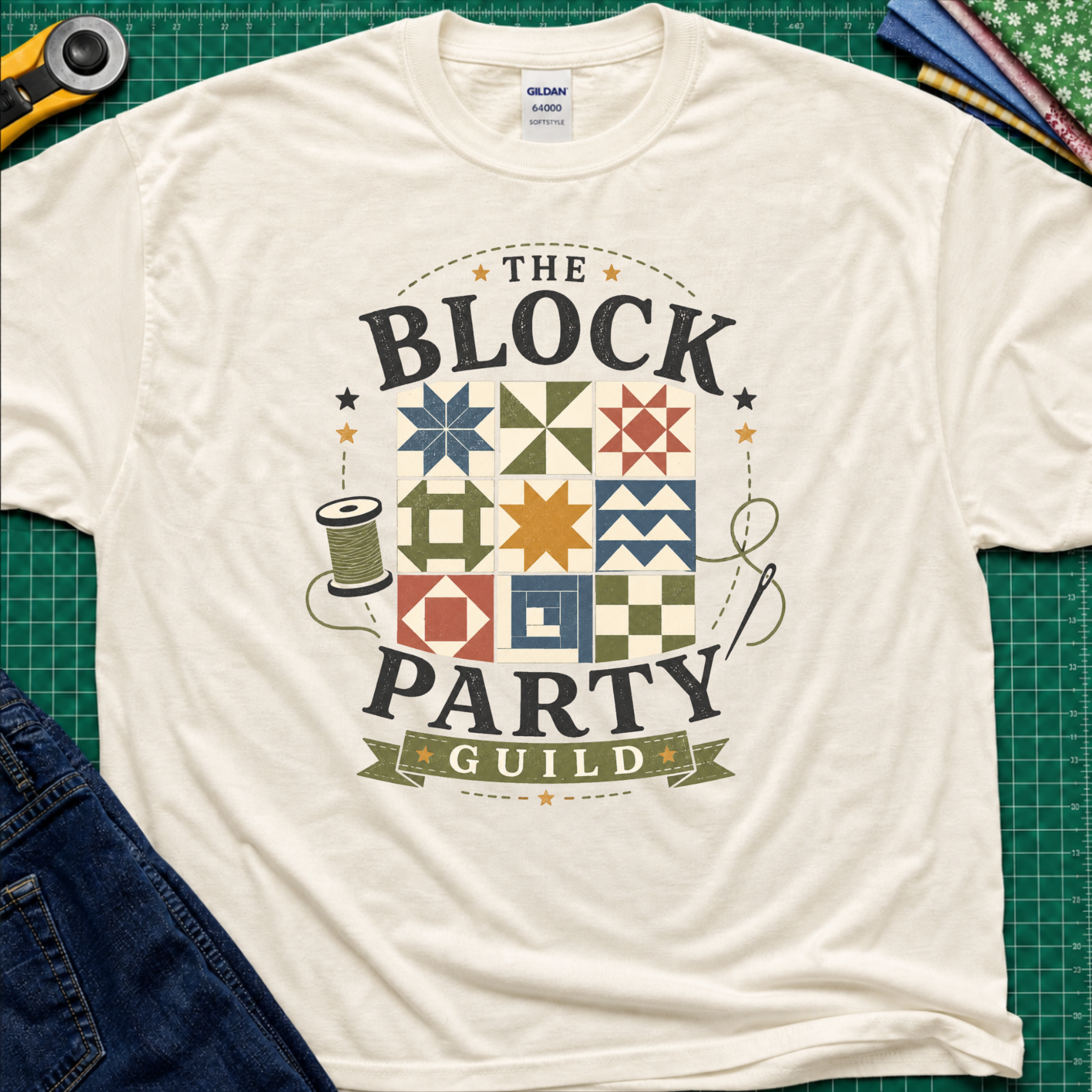 A light cream Gildan t-shirt with a vintage-style graphic reading 'THE BLOCK PARTY GUILD' around a set of six quilt blocks. The shirt is laid flat on a green cutting mat with sewing-themed props like a rotary cutter, fabric, and jeans partially visible in the corners.