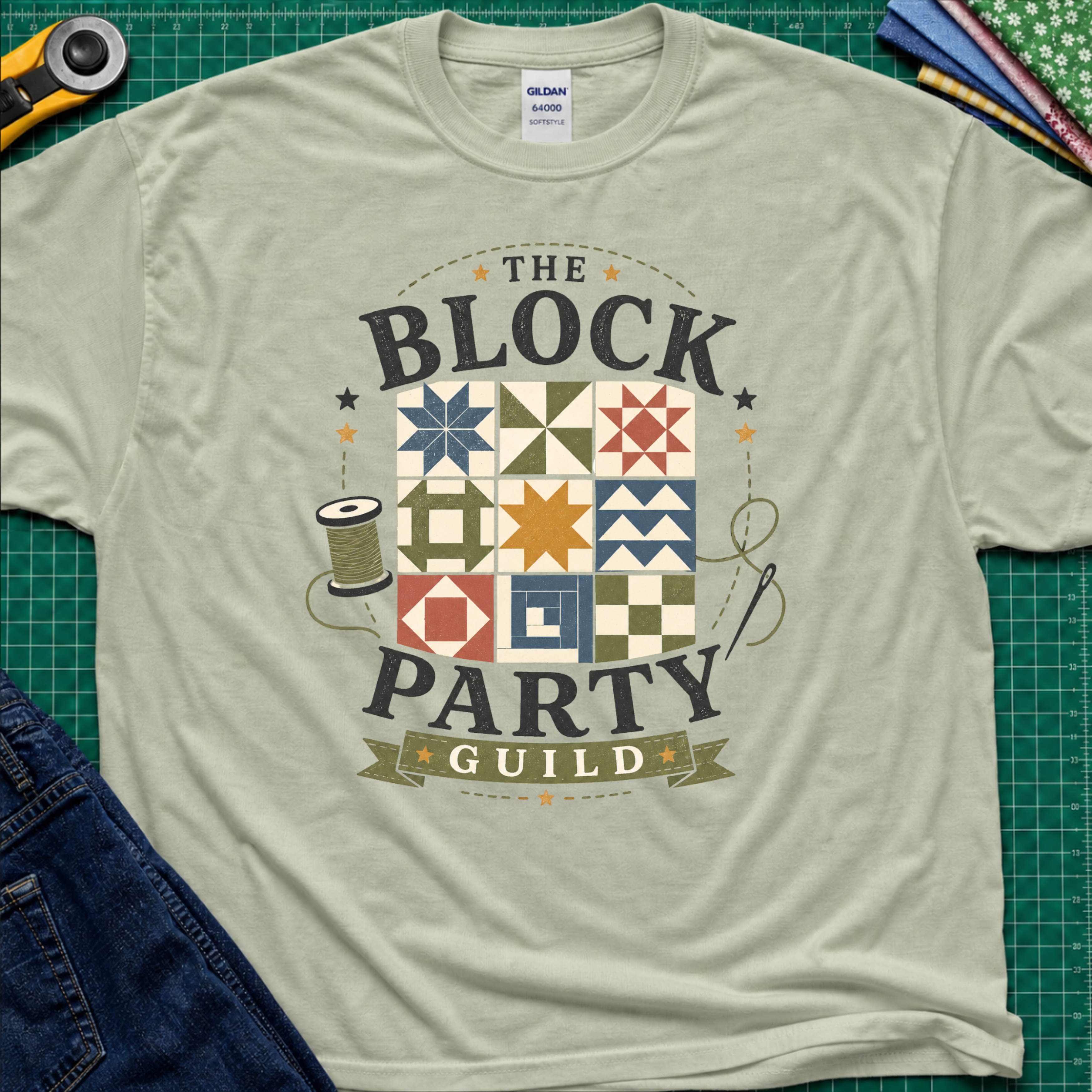 A light sage green Gildan t-shirt with a vintage-style graphic reading 'THE BLOCK PARTY GUILD' around a set of six quilt blocks. The shirt is laid flat on a green cutting mat with sewing-themed props like a rotary cutter, fabric, and jeans partially visible in the corners.