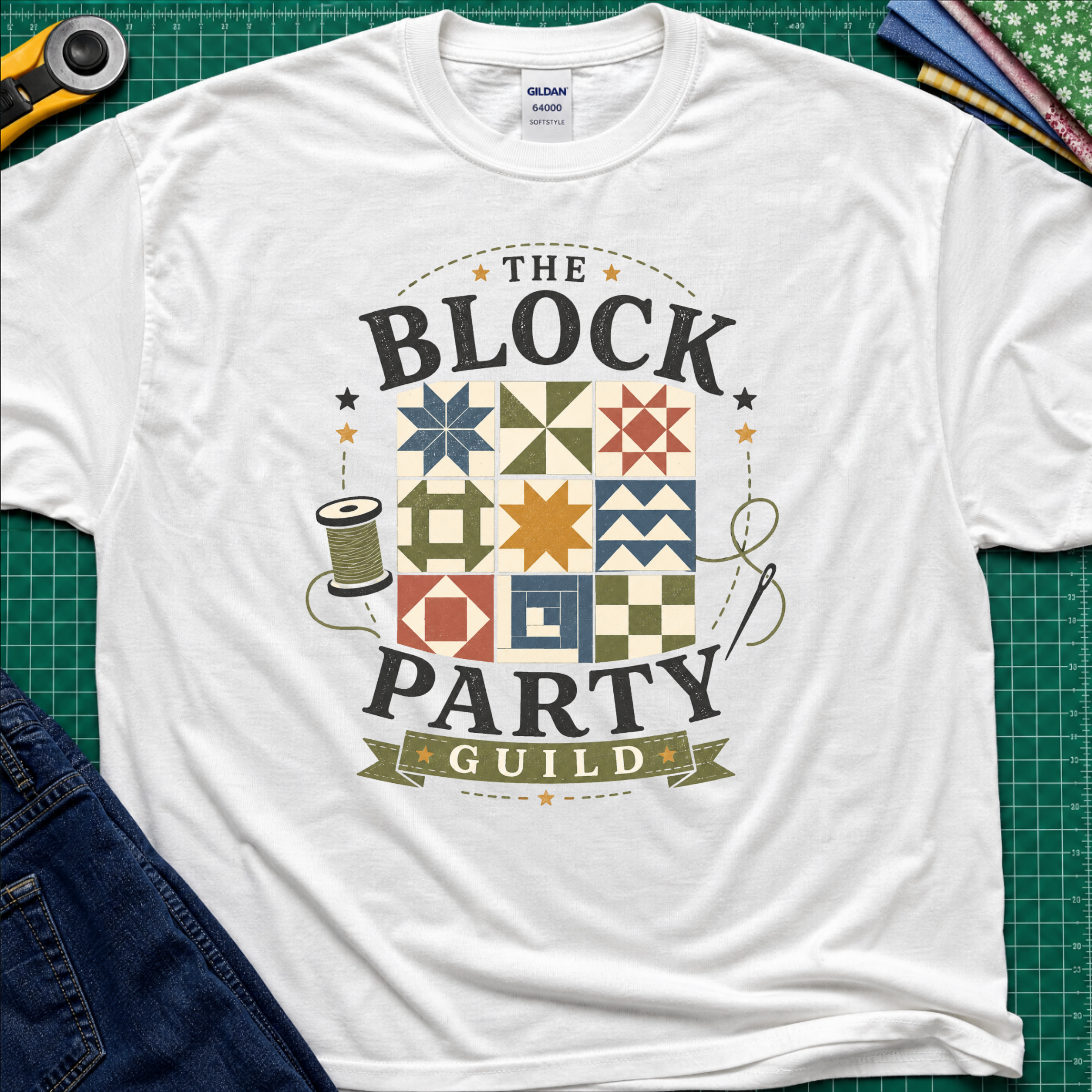 A white Gildan t-shirt with a vintage-style graphic reading 'THE BLOCK PARTY GUILD' around a set of six quilt blocks. The shirt is laid flat on a green cutting mat with sewing-themed props like a rotary cutter, fabric, and jeans partially visible in the corners.