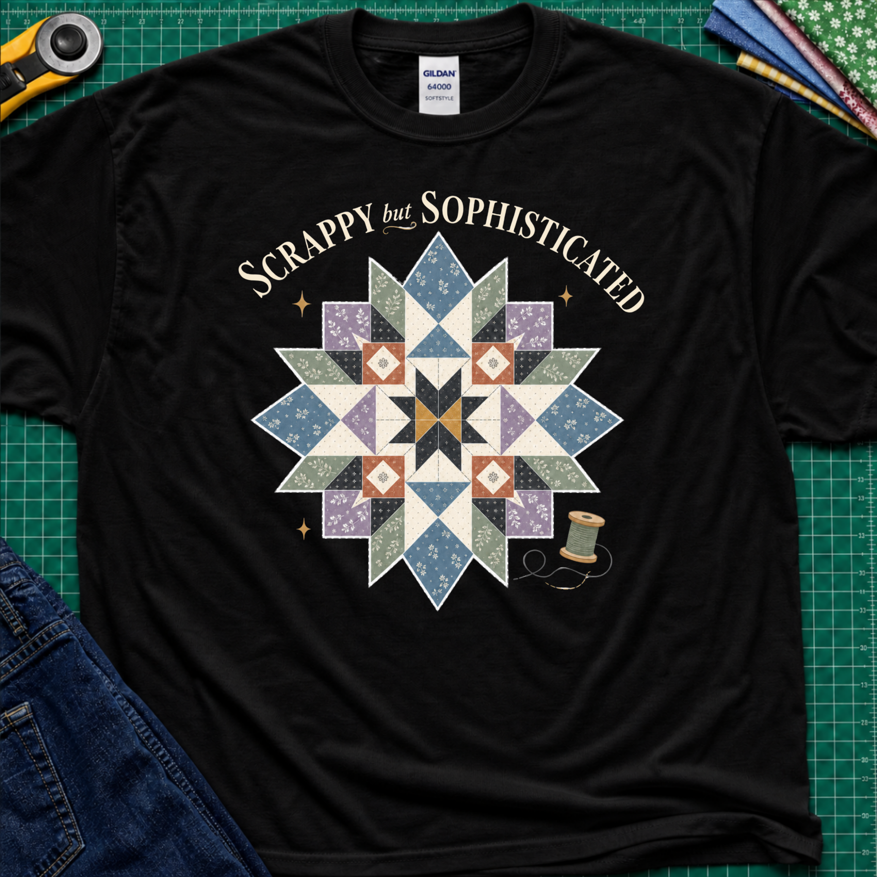 A black Gildan t-shirt with a colorful quilted starburst graphic and the text 'Scrappy but Sophisticated' printed on the front, styled on a green cutting mat with sewing tools and denim.
