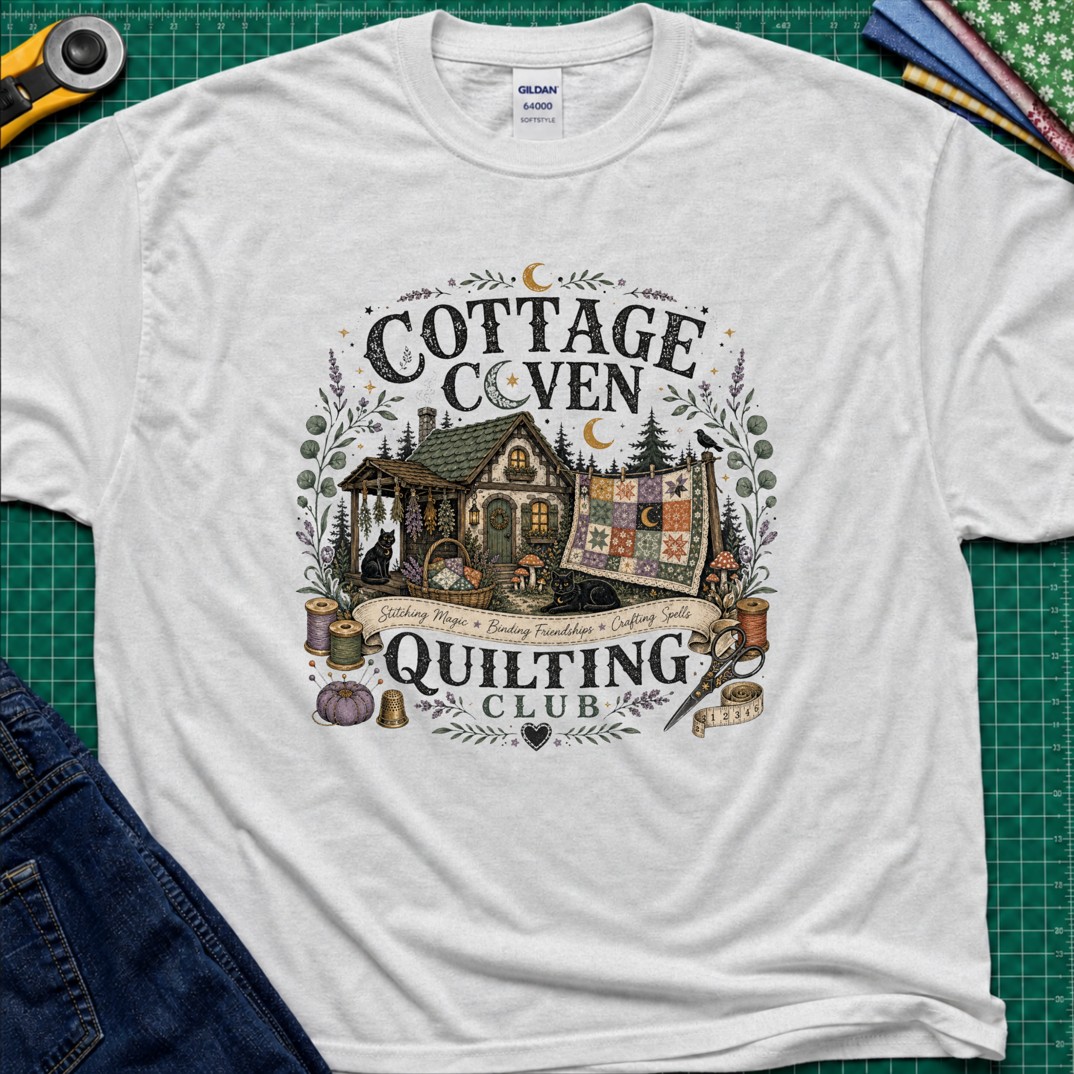 A light grey Gildan t-shirt laid flat on a green cutting mat, featuring a detailed graphic of a cottage and a quilt with the text 'COTTAGE COVEN QUILTING CLUB'. The shirt is styled with a rotary cutter, denim jeans, and other crafting tools.