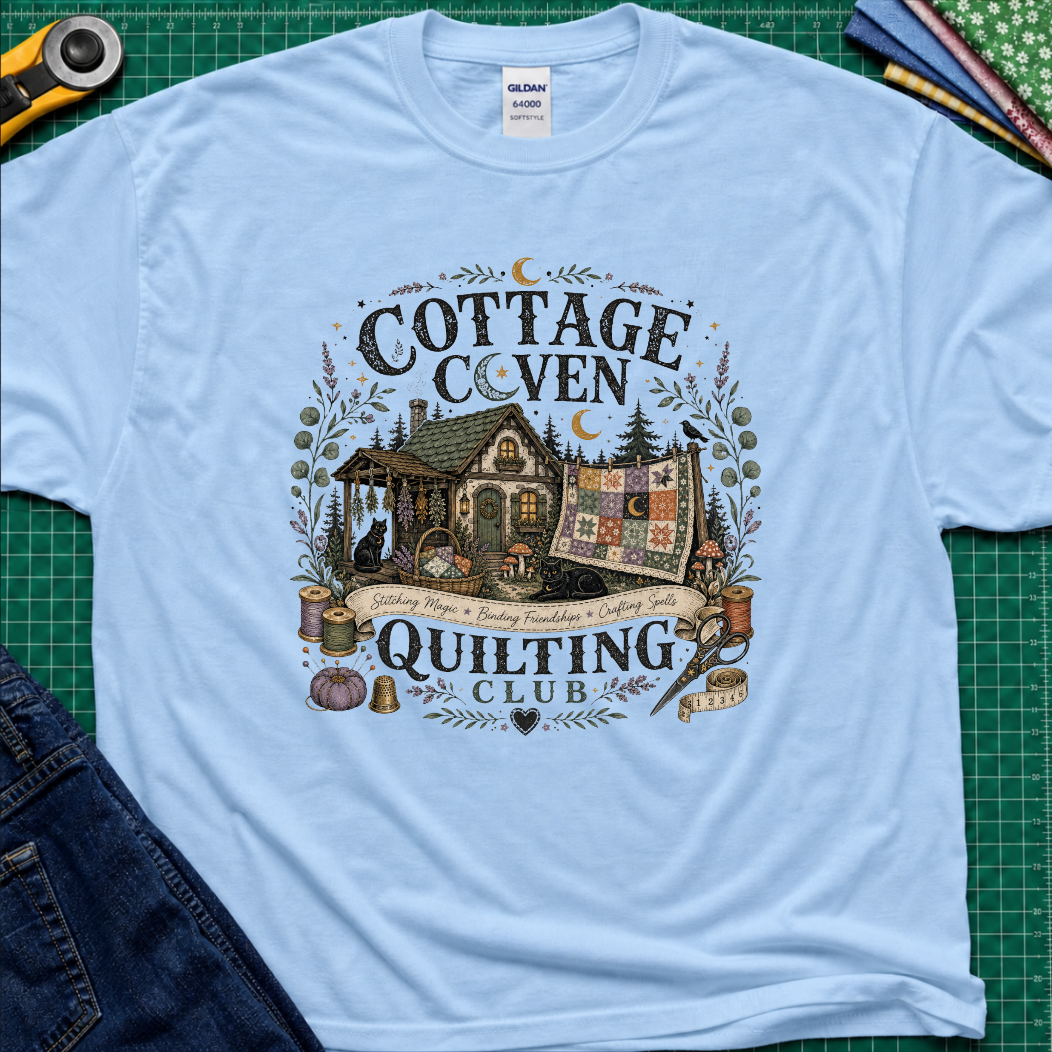 A light blue Gildan t-shirt laid flat on a green cutting mat, featuring a detailed graphic of a cottage and a quilt with the text 'COTTAGE COVEN QUILTING CLUB'. The shirt is styled with a rotary cutter, denim jeans, and other crafting tools.