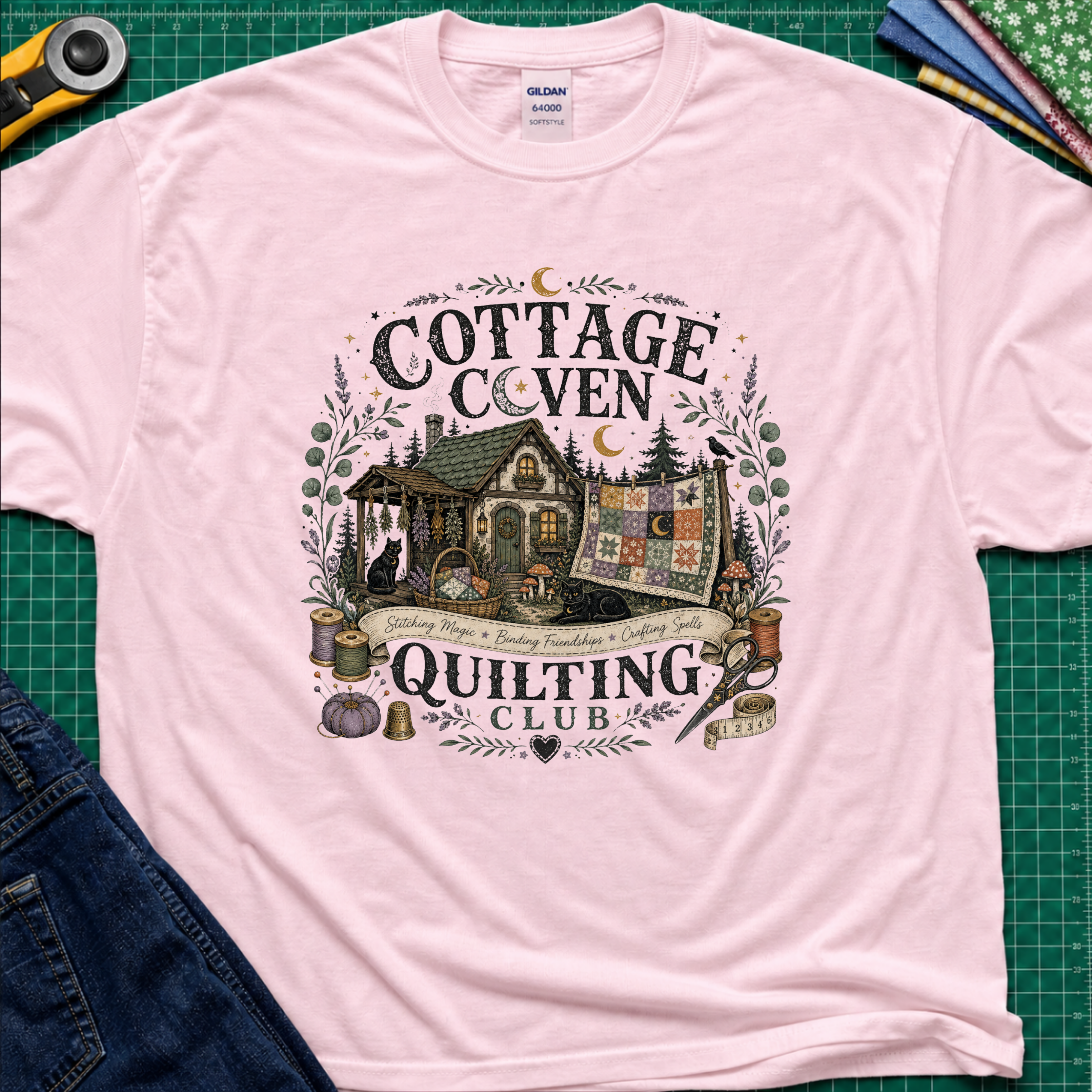 A light pink Gildan t-shirt laid flat on a green cutting mat, featuring a detailed graphic of a cottage and a quilt with the text 'COTTAGE COVEN QUILTING CLUB'. The shirt is styled with a rotary cutter, denim jeans, and other crafting tools.