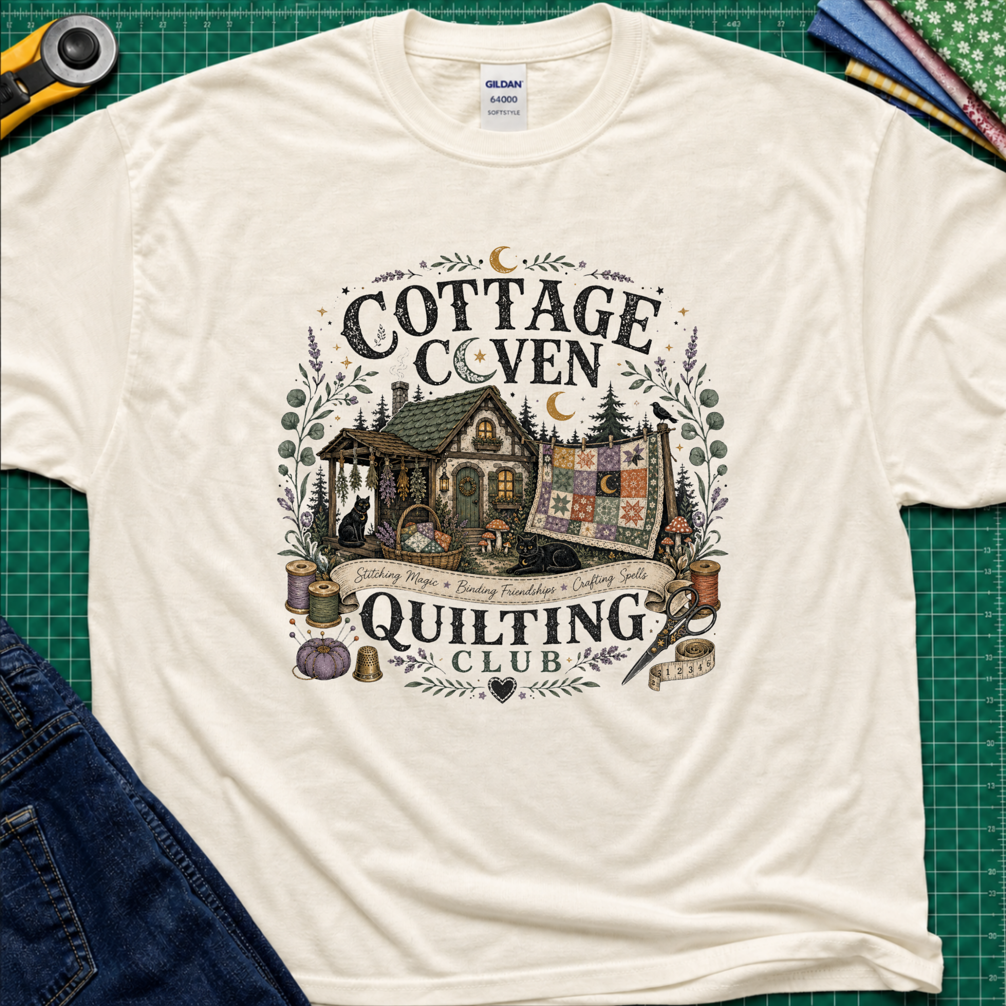 A light cream Gildan t-shirt laid flat on a green cutting mat, featuring a detailed graphic of a cottage and a quilt with the text 'COTTAGE COVEN QUILTING CLUB'. The shirt is styled with a rotary cutter, denim jeans, and other crafting tools.