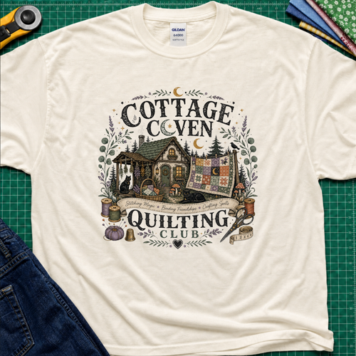 A light cream Gildan t-shirt laid flat on a green cutting mat, featuring a detailed graphic of a cottage and a quilt with the text 'COTTAGE COVEN QUILTING CLUB'. The shirt is styled with a rotary cutter, denim jeans, and other crafting tools.