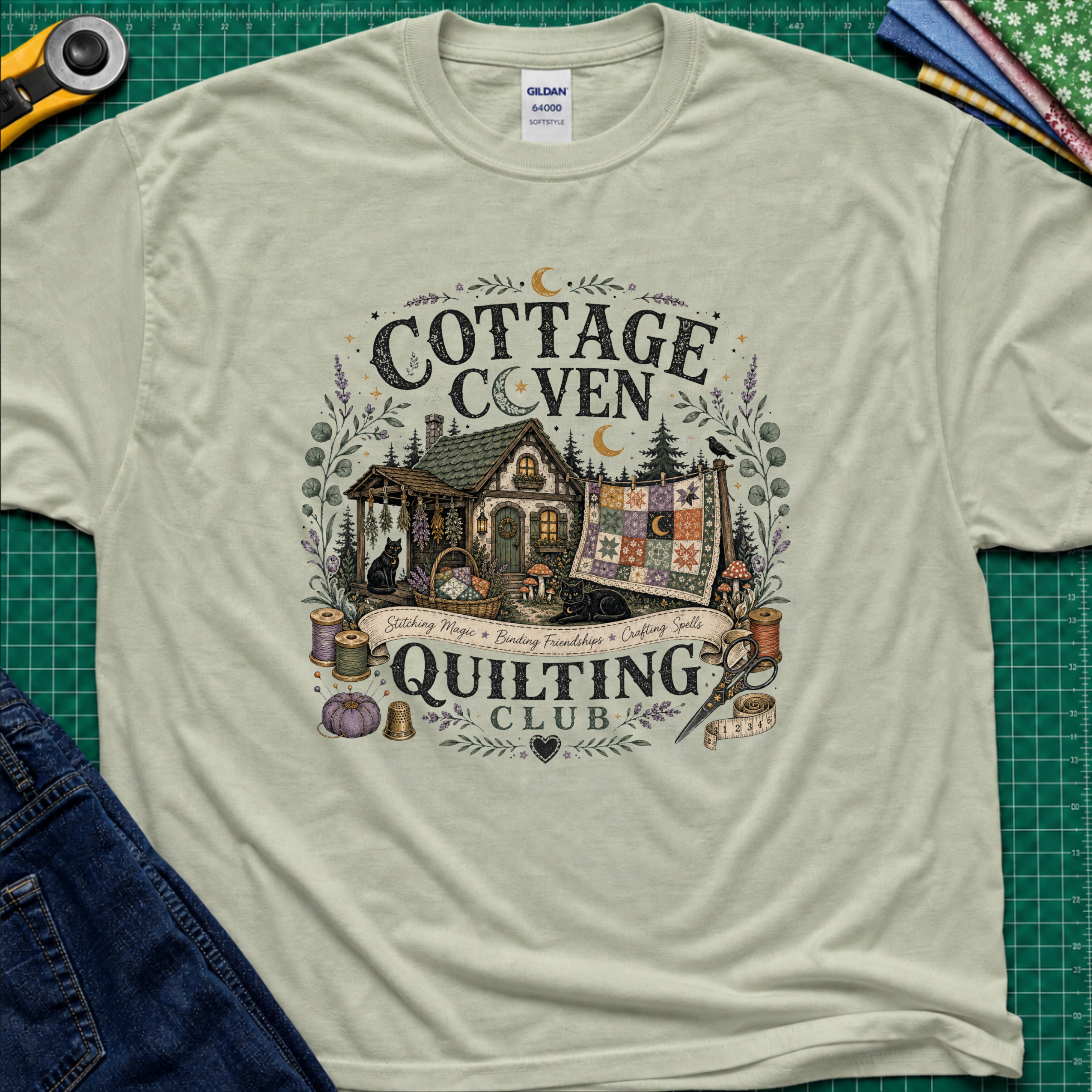 A light sage green Gildan t-shirt laid flat on a green cutting mat, featuring a detailed graphic of a cottage and a quilt with the text 'COTTAGE COVEN QUILTING CLUB'. The shirt is styled with a rotary cutter, denim jeans, and other crafting tools.
