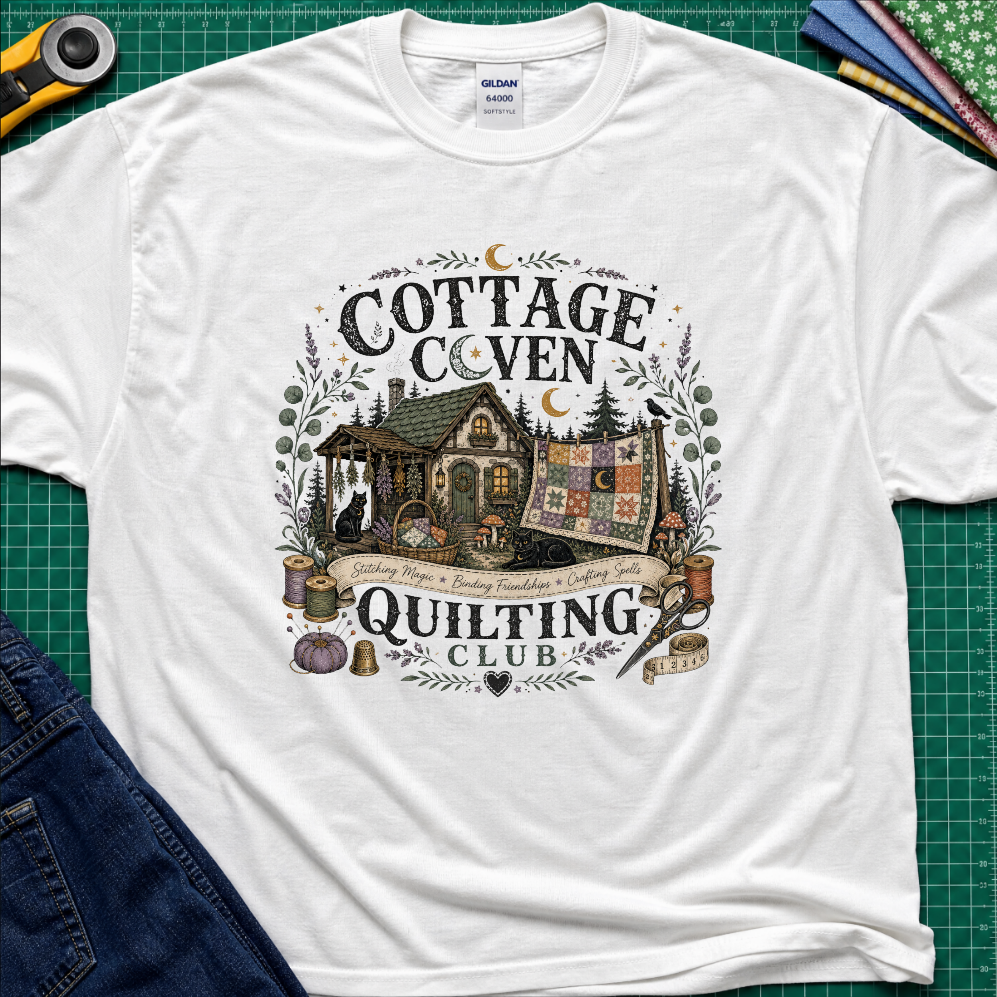 A white Gildan t-shirt laid flat on a green cutting mat, featuring a detailed graphic of a cottage and a quilt with the text 'COTTAGE COVEN QUILTING CLUB'. The shirt is styled with a rotary cutter, denim jeans, and other crafting tools.