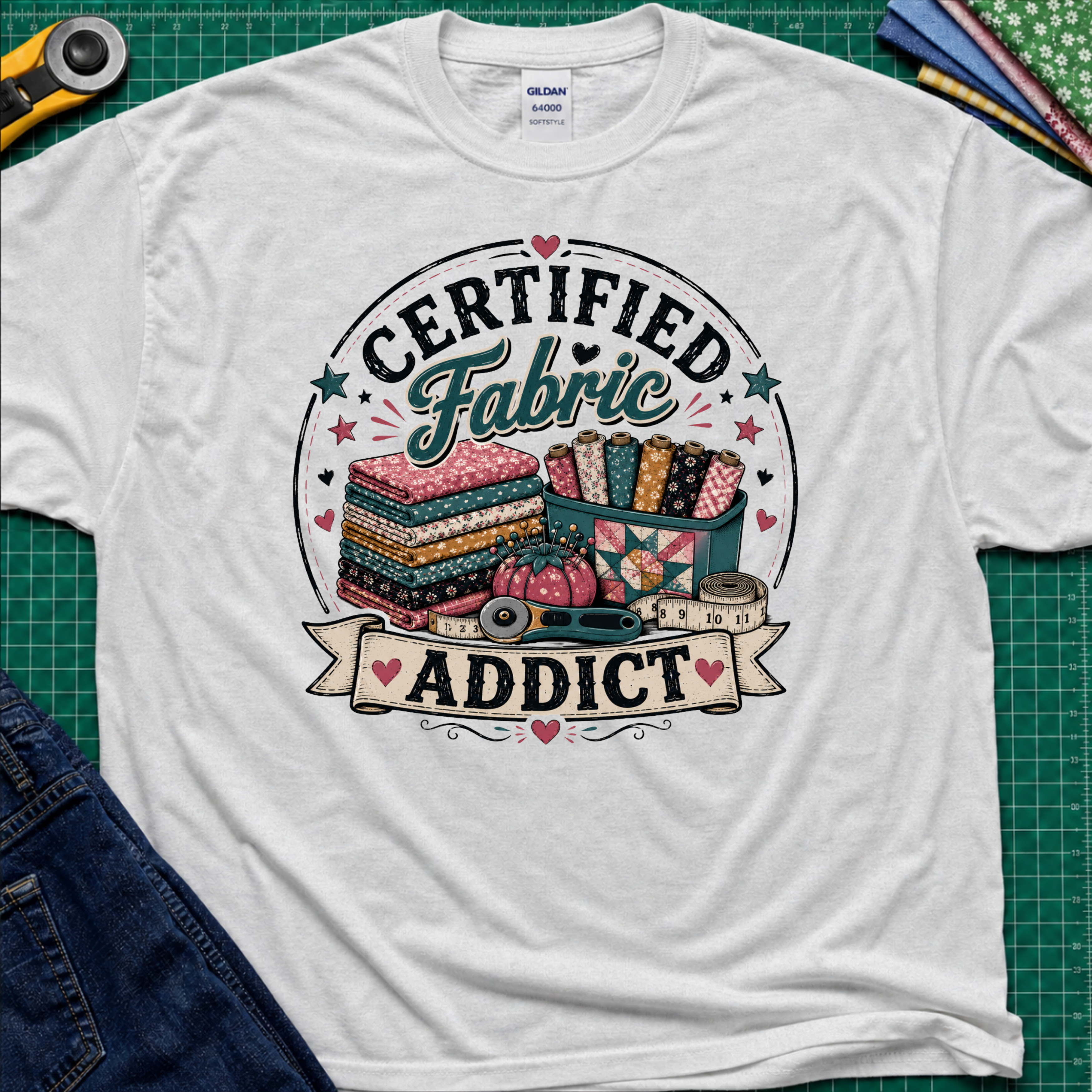 A light ice grey Gildan t-shirt featuring a colorful graphic that reads 'CERTIFIED Fabric ADDICT' with illustrations of stacked fabric bolts, a pin cusion, measuring tape and rotary cutter. The shirt lays flat on a green cutting mat with denim jeans, and sewing supplies partially visible around the shirt.