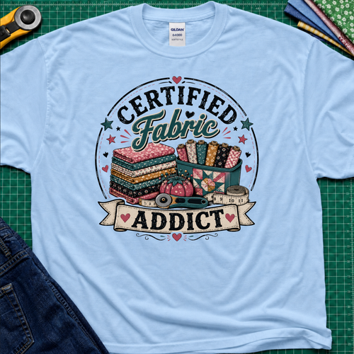 A light blue Gildan t-shirt featuring a colorful graphic that reads 'CERTIFIED Fabric ADDICT' with illustrations of stacked fabric bolts, a pin cusion, measuring tape and rotary cutter. The shirt lays flat on a green cutting mat with denim jeans, and sewing supplies partially visible around the shirt.