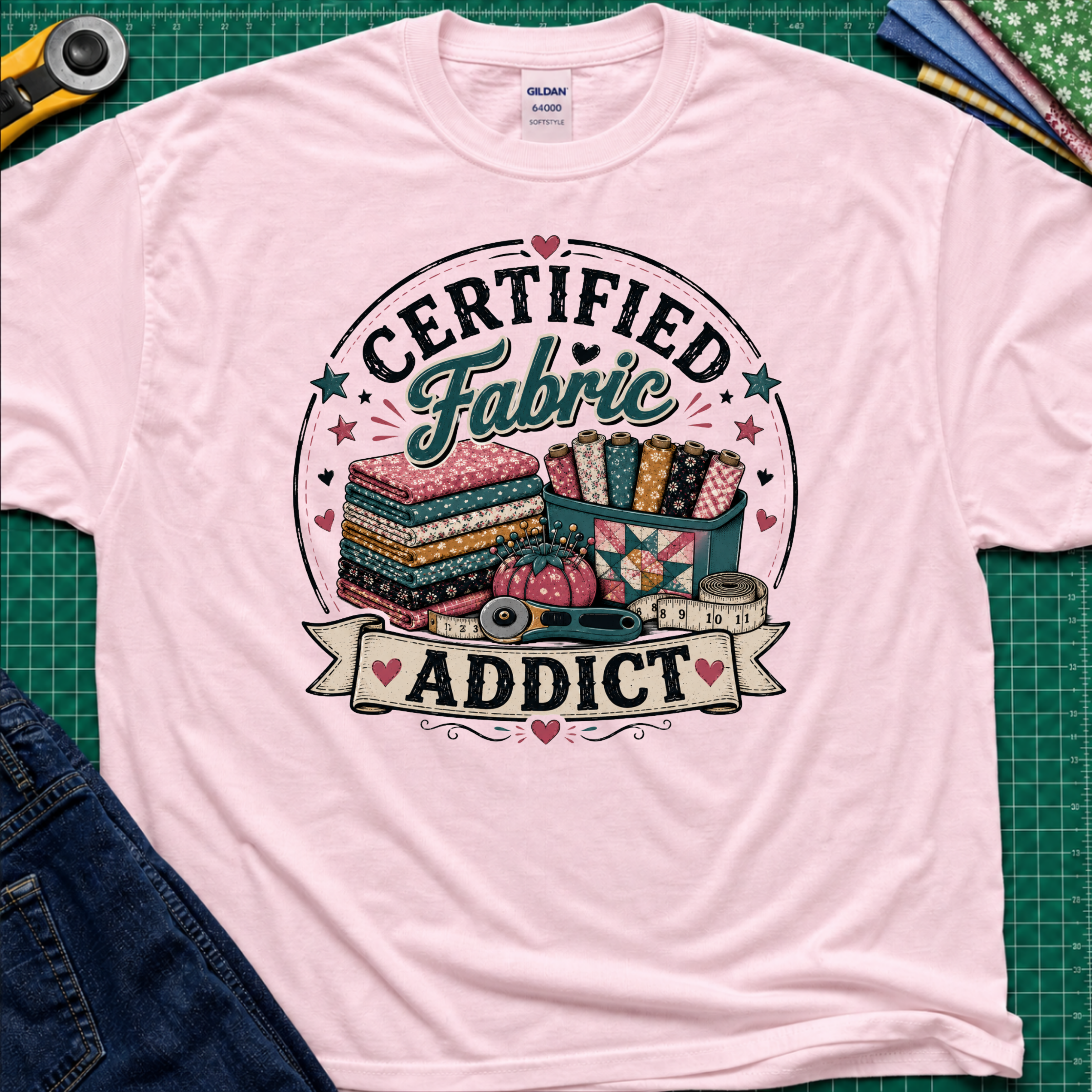 A light pink Gildan t-shirt featuring a colorful graphic that reads 'CERTIFIED Fabric ADDICT' with illustrations of stacked fabric bolts, a pin cusion, measuring tape and rotary cutter. The shirt lays flat on a green cutting mat with denim jeans, and sewing supplies partially visible around the shirt.