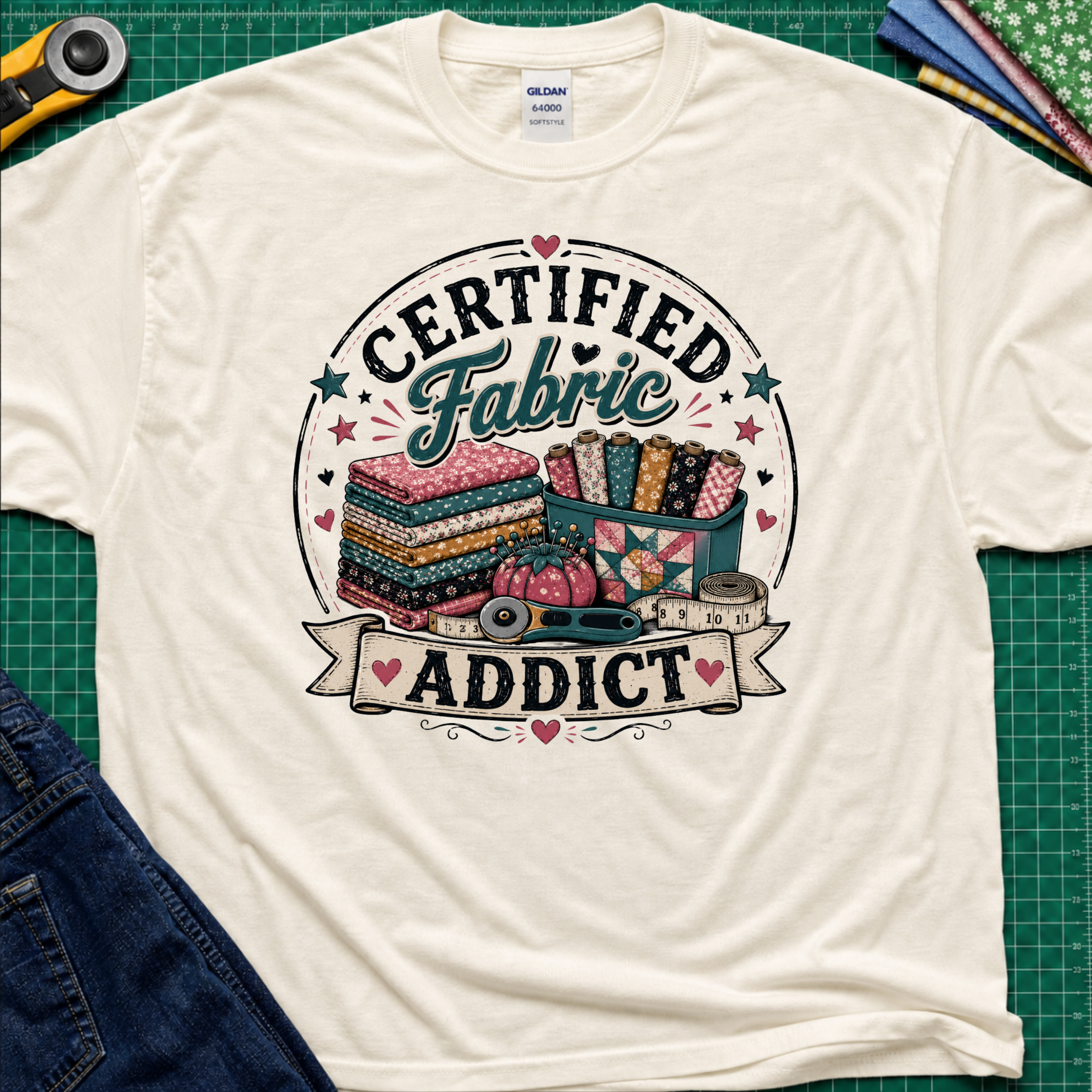 A light cream Gildan t-shirt featuring a colorful graphic that reads 'CERTIFIED Fabric ADDICT' with illustrations of stacked fabric bolts, a pin cusion, measuring tape and rotary cutter. The shirt lays flat on a green cutting mat with denim jeans, and sewing supplies partially visible around the shirt.