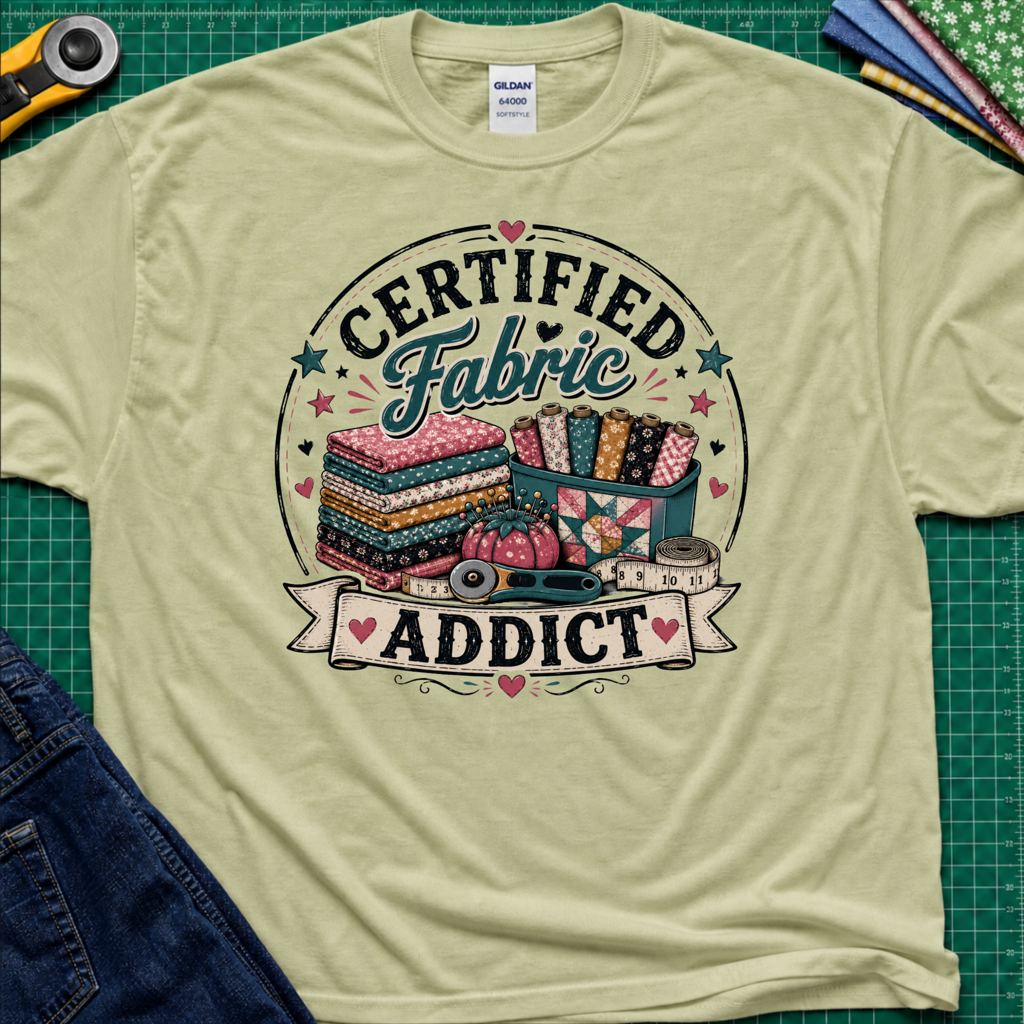 A light pistachio green Gildan t-shirt featuring a colorful graphic that reads 'CERTIFIED Fabric ADDICT' with illustrations of stacked fabric bolts, a pin cusion, measuring tape and rotary cutter. The shirt lays flat on a green cutting mat with denim jeans, and sewing supplies partially visible around the shirt.