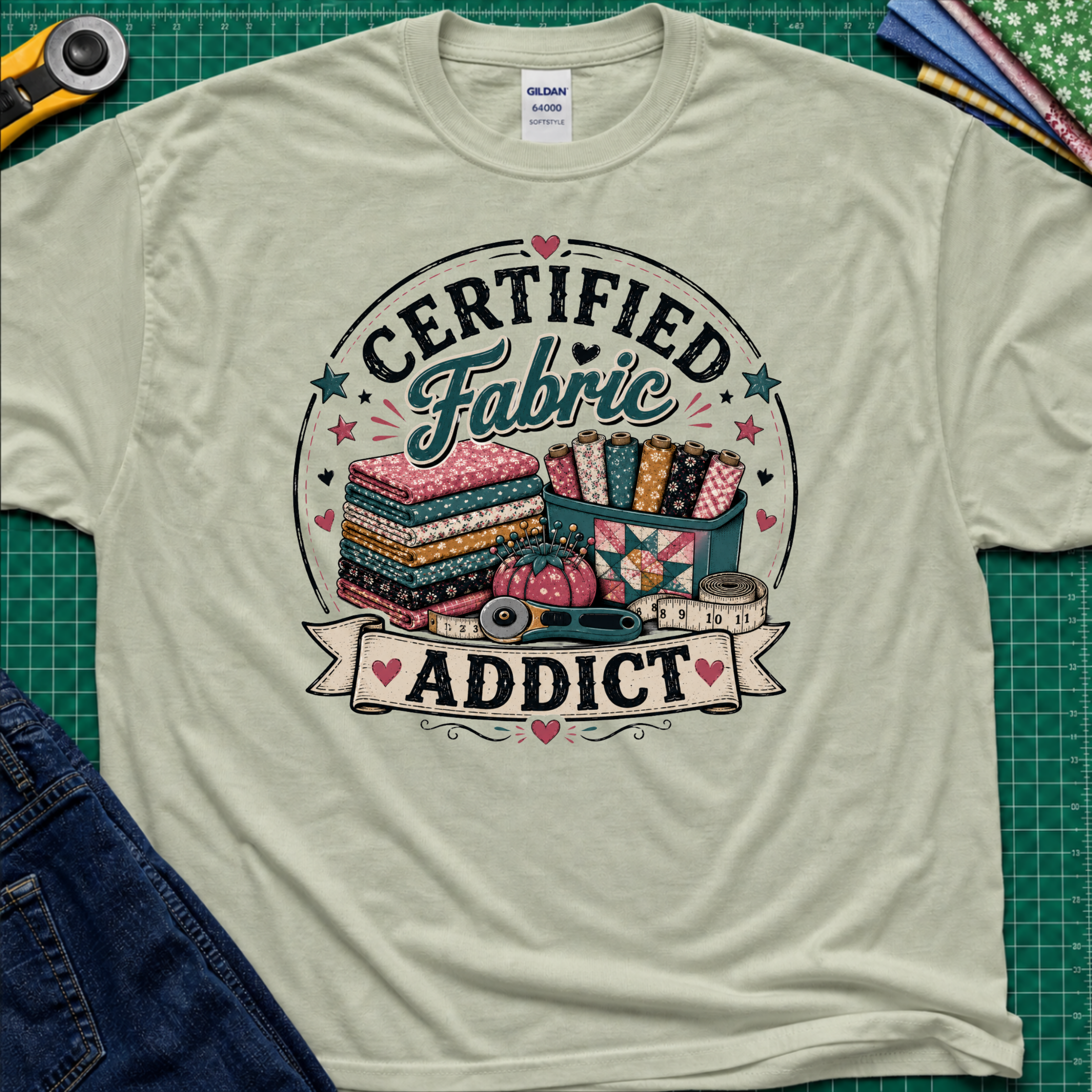A light sage green Gildan t-shirt featuring a colorful graphic that reads 'CERTIFIED Fabric ADDICT' with illustrations of stacked fabric bolts, a pin cusion, measuring tape and rotary cutter. The shirt lays flat on a green cutting mat with denim jeans, and sewing supplies partially visible around the shirt.