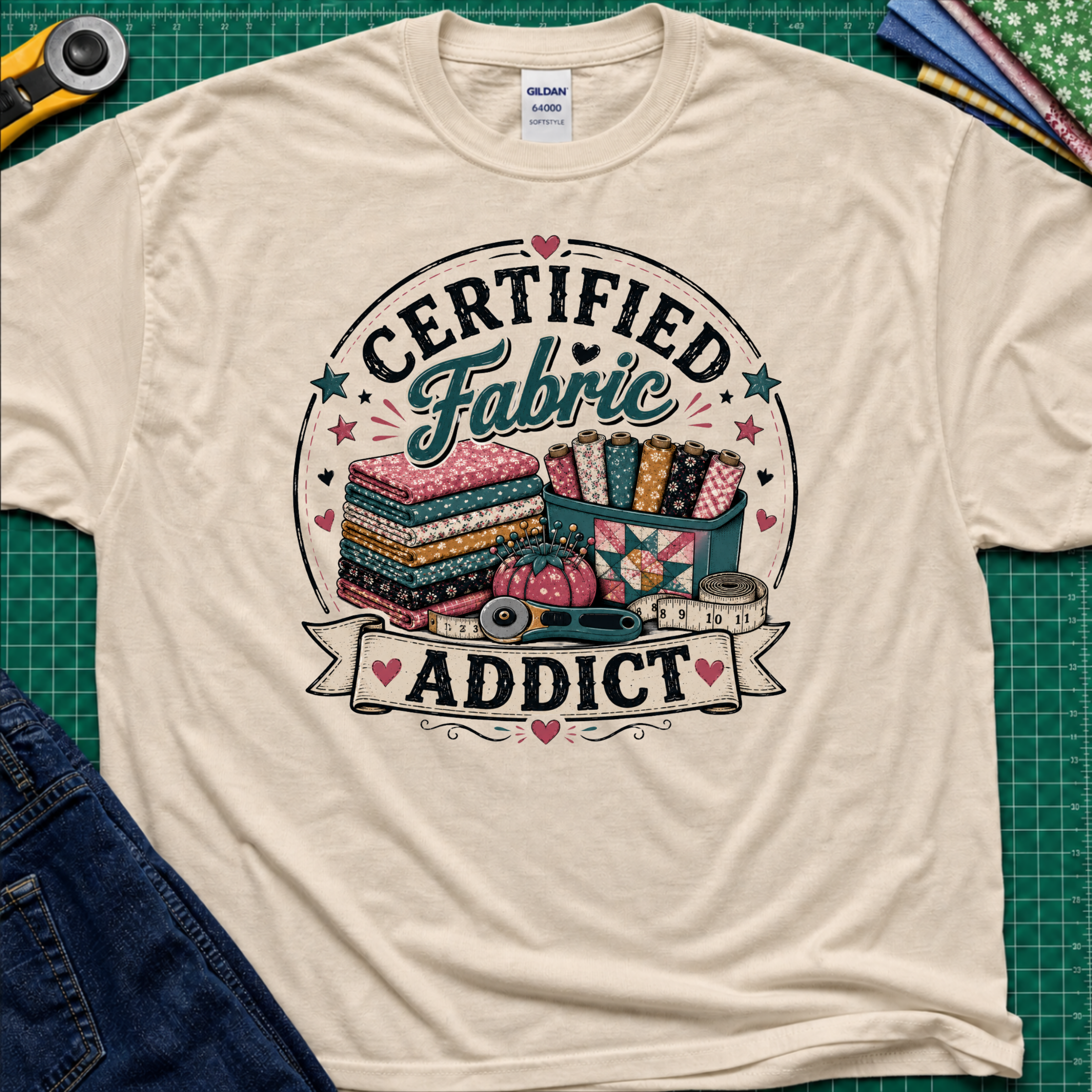 A dark cream Gildan t-shirt featuring a colorful graphic that reads 'CERTIFIED Fabric ADDICT' with illustrations of stacked fabric bolts, a pin cusion, measuring tape and rotary cutter. The shirt lays flat on a green cutting mat with denim jeans, and sewing supplies partially visible around the shirt.
