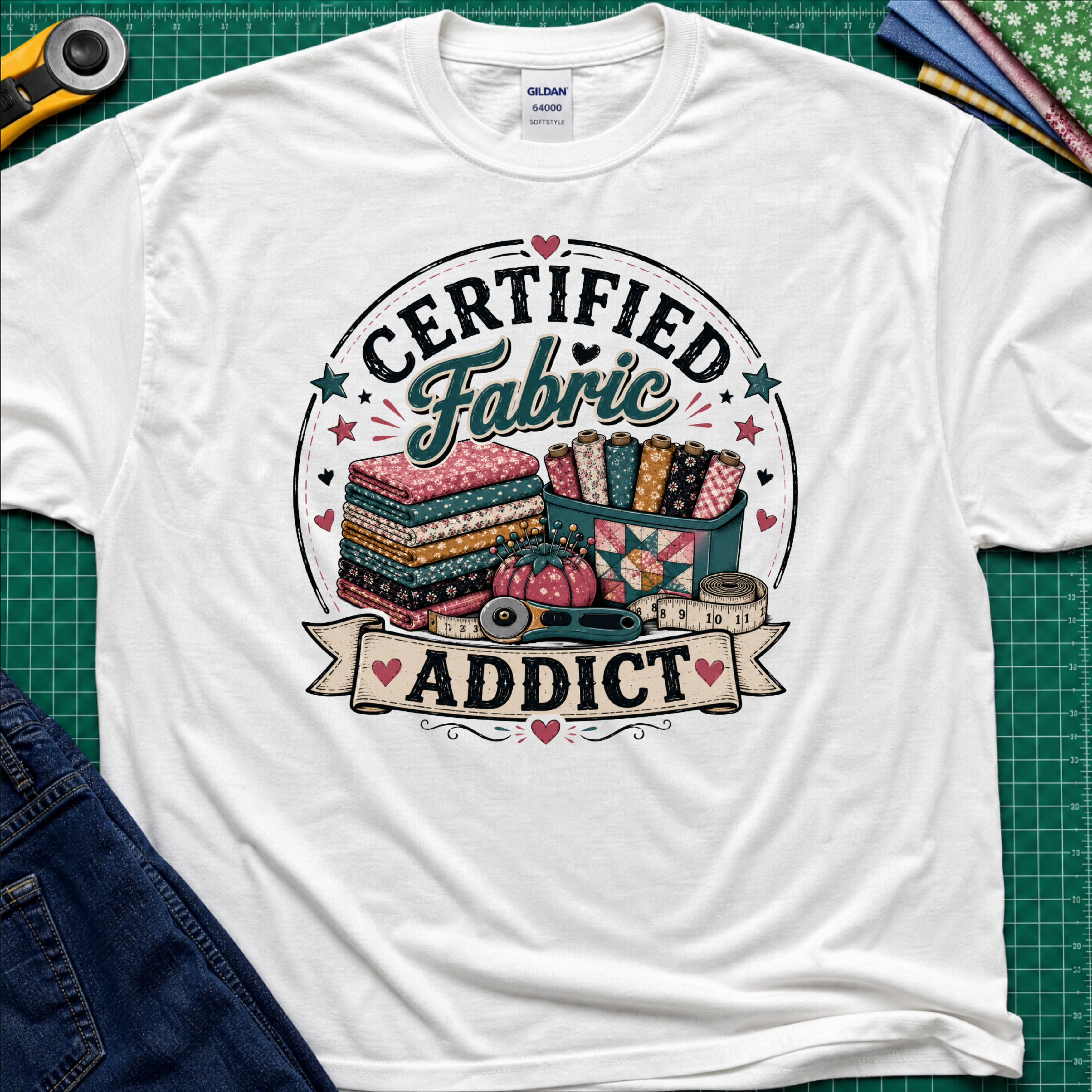 A white grey Gildan t-shirt featuring a colorful graphic that reads 'CERTIFIED Fabric ADDICT' with illustrations of stacked fabric bolts, a pin cusion, measuring tape and rotary cutter. The shirt lays flat on a green cutting mat with denim jeans, and sewing supplies partially visible around the shirt.
