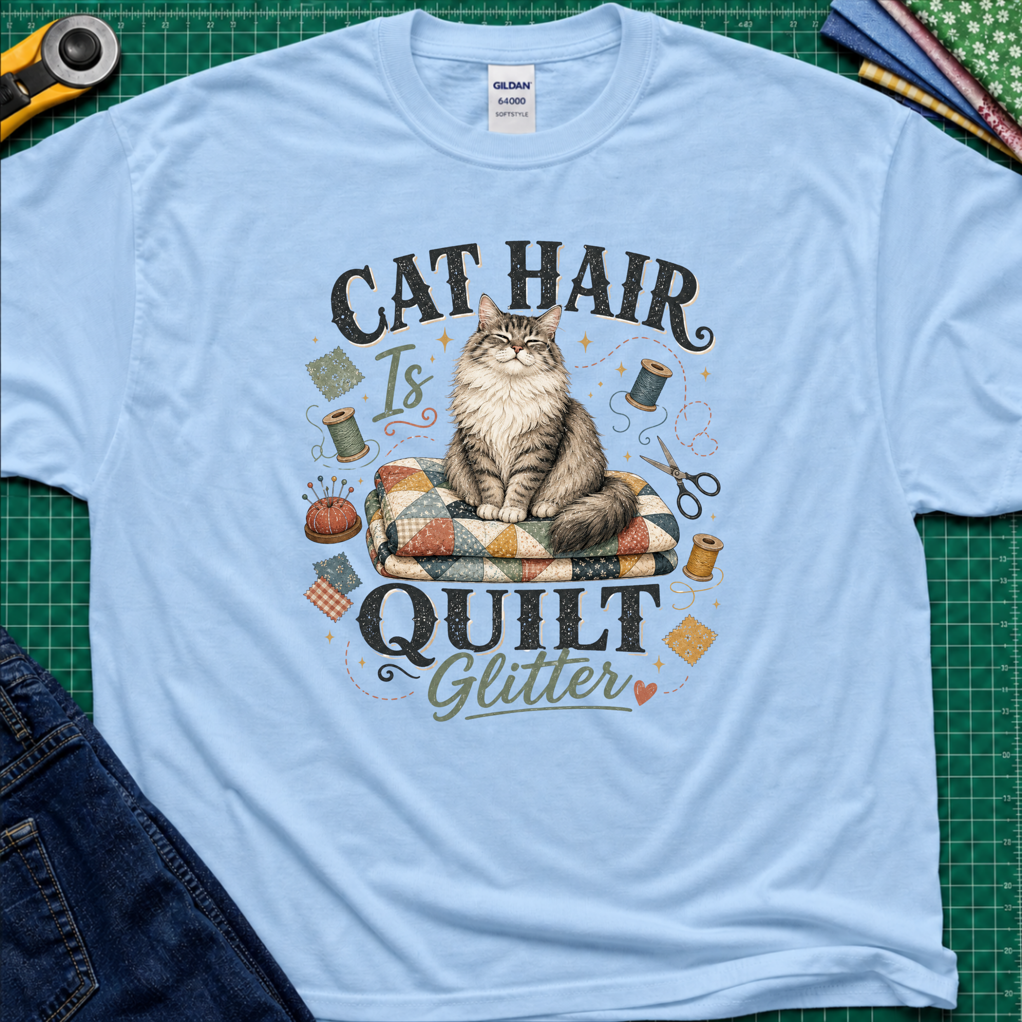A light blue Gildan t-shirt laid flat on a green cutting mat, featuring a graphic of a fluffy cat sitting on a quilt with the text 'CAT HAIR Is QUILT Glitter' surrounded by sewing-themed illustrations.