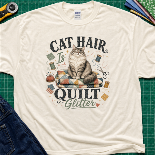 A light cream Gildan t-shirt laid flat on a green cutting mat, featuring a graphic of a fluffy cat sitting on a quilt with the text 'CAT HAIR Is QUILT Glitter' surrounded by sewing-themed illustrations.