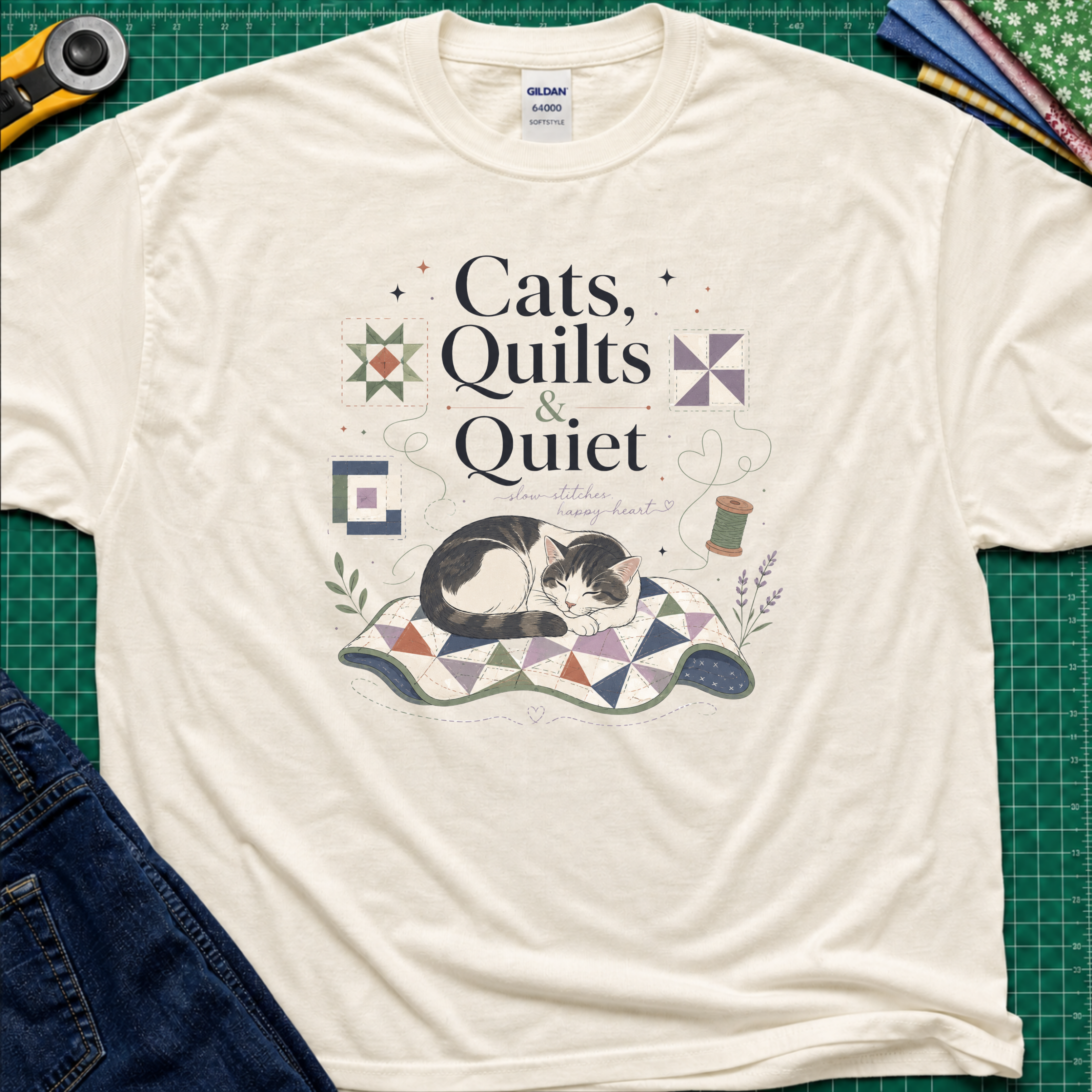 A light cream Gildant-shirt featuring a cat laying on a quilt and the text 'Cats, Quilts & Quiet', with small quilt blocks and thread.  The shirt lays on a green cutting mat with rotary cutter, jeans, and fabric stack partially visible around the shirt.