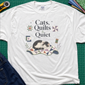 A white Gildan t-shirt featuring a cat laying on a quilt and the text 'Cats, Quilts & Quiet', with small quilt blocks and thread. The shirt lays on a green cutting mat with rotary cutter, jeans, and fabric stack partially visible around the shirt.
