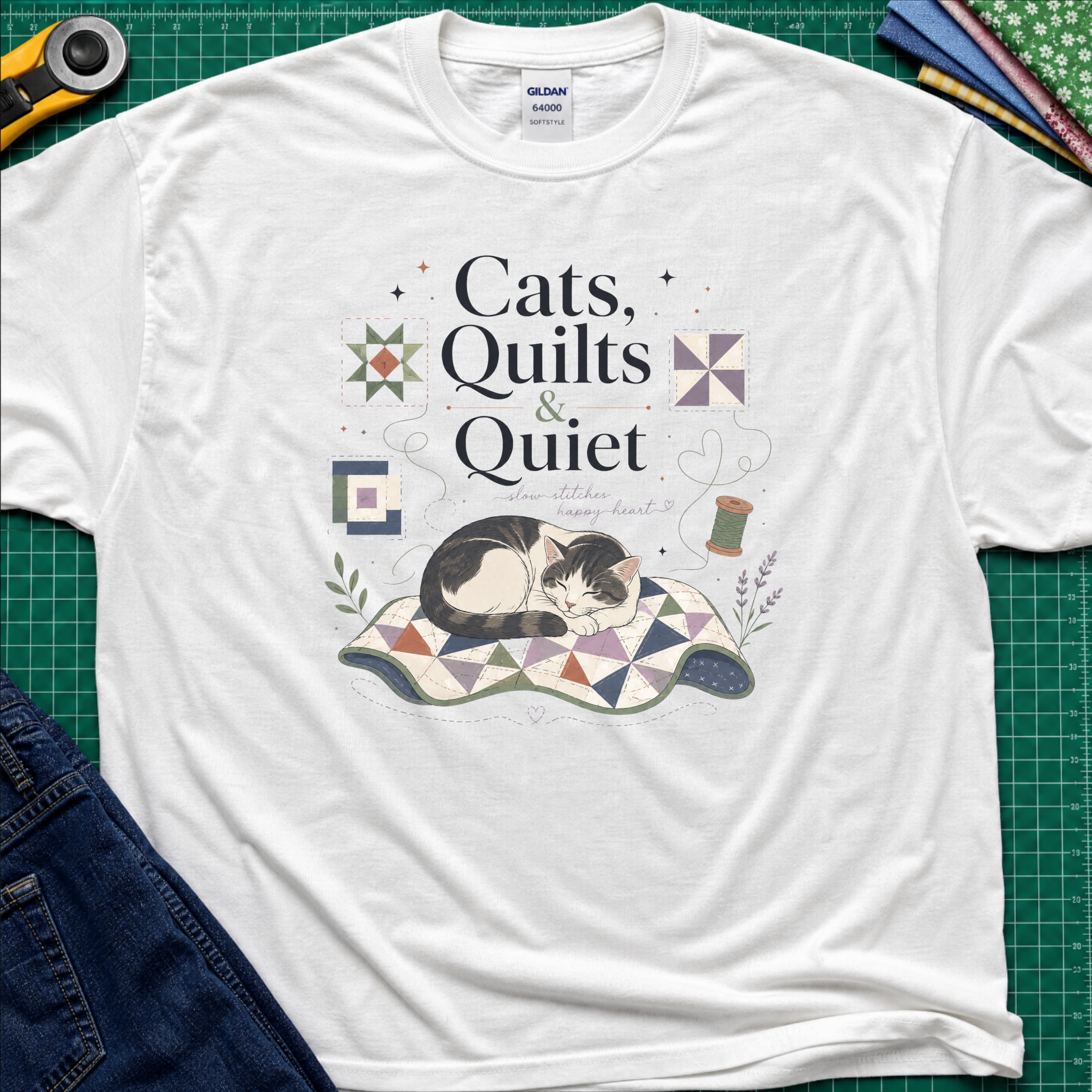 A white Gildan t-shirt featuring a cat laying on a quilt and the text 'Cats, Quilts & Quiet', with small quilt blocks and thread. The shirt lays on a green cutting mat with rotary cutter, jeans, and fabric stack partially visible around the shirt.