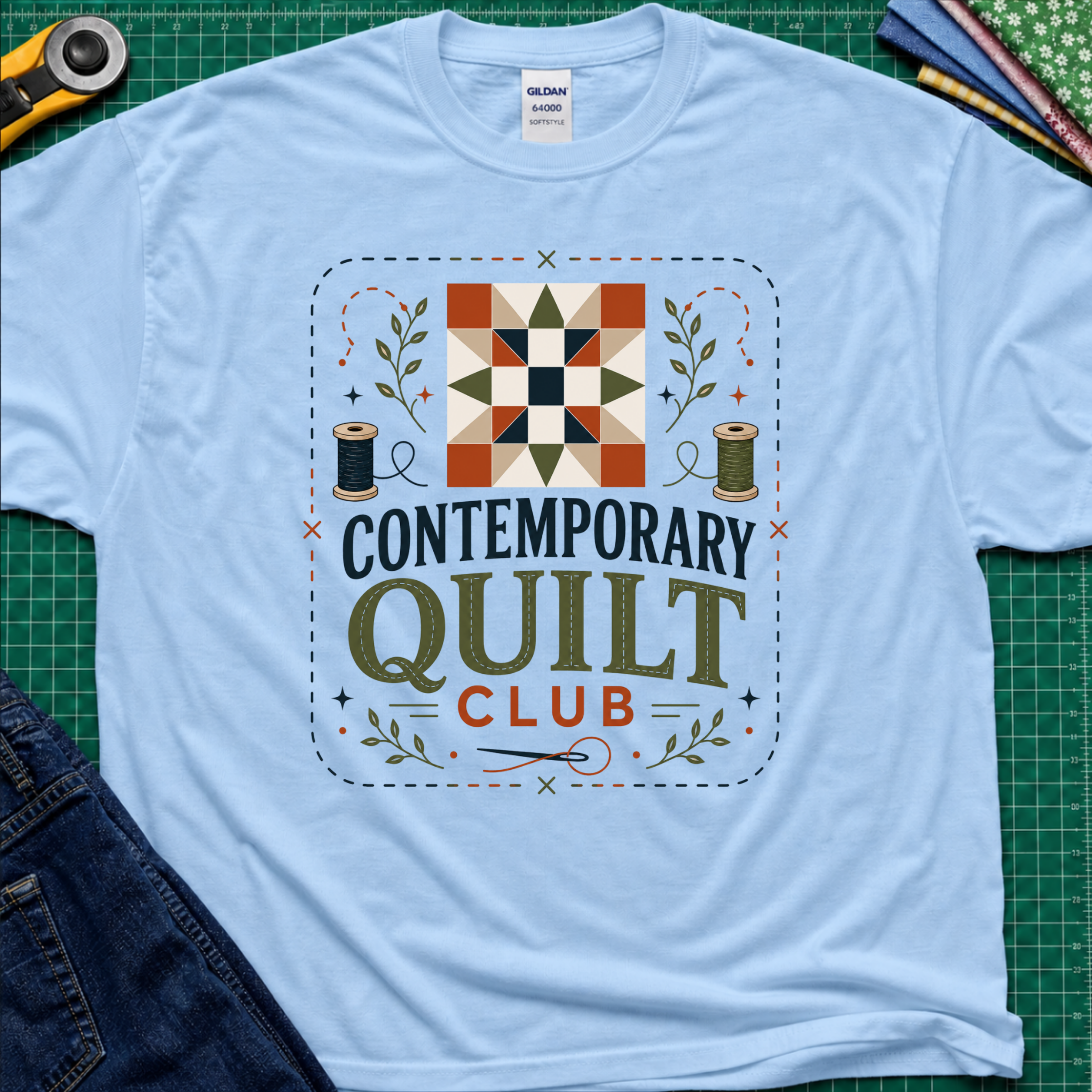 A light blue Gildan t-shirt laid flat on a green cutting mat, featuring a retro-style 'Contemporary Quilt Club' graphic with a quilt pattern, spools of thread, and decorative elements.