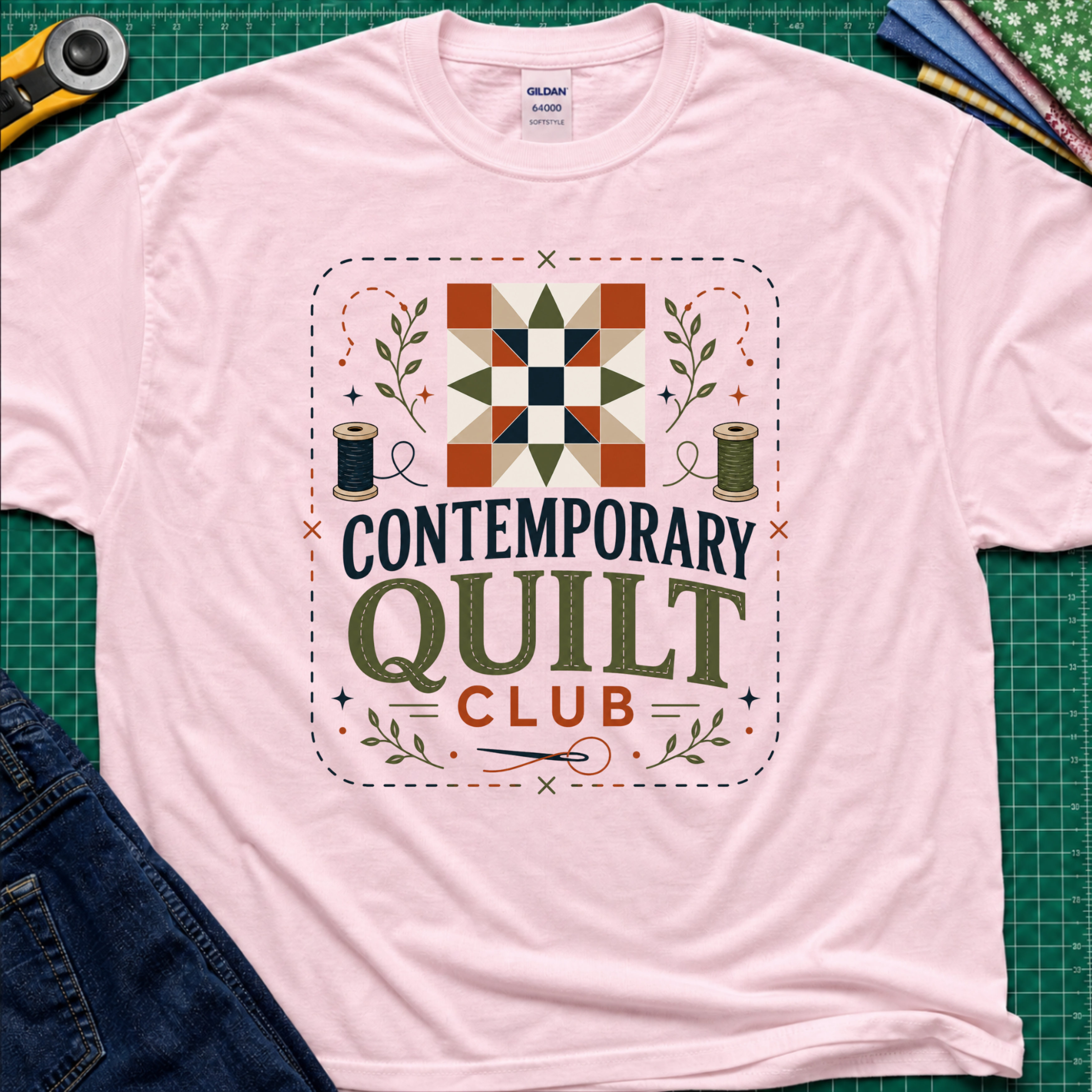 A light pink Gildan t-shirt laid flat on a green cutting mat, featuring a retro-style 'Contemporary Quilt Club' graphic with a quilt pattern, spools of thread, and decorative elements.