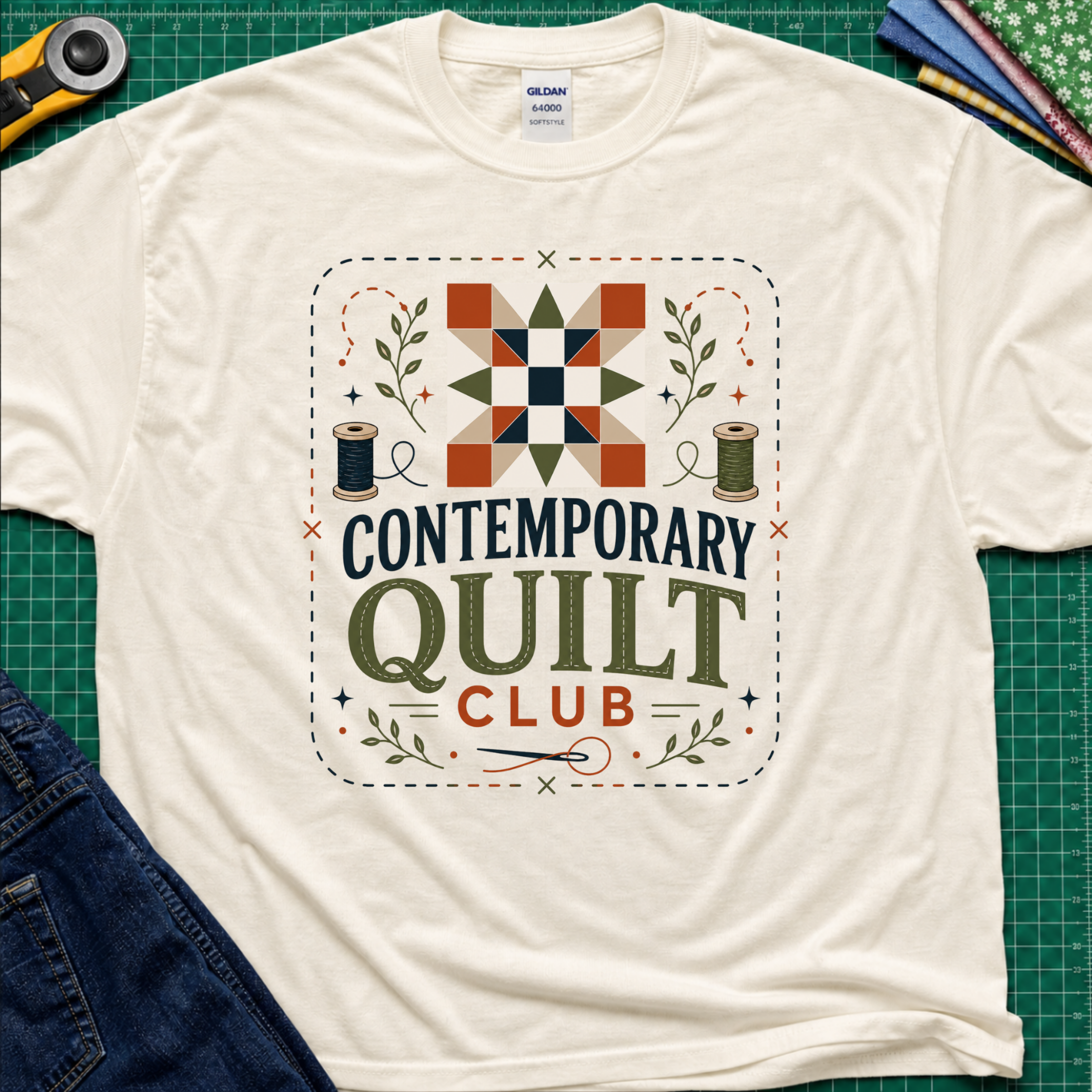 A light cream Gildan t-shirt laid flat on a green cutting mat, featuring a retro-style 'Contemporary Quilt Club' graphic with a quilt pattern, spools of thread, and decorative elements.