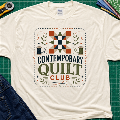 A light cream Gildan t-shirt laid flat on a green cutting mat, featuring a retro-style 'Contemporary Quilt Club' graphic with a quilt pattern, spools of thread, and decorative elements.