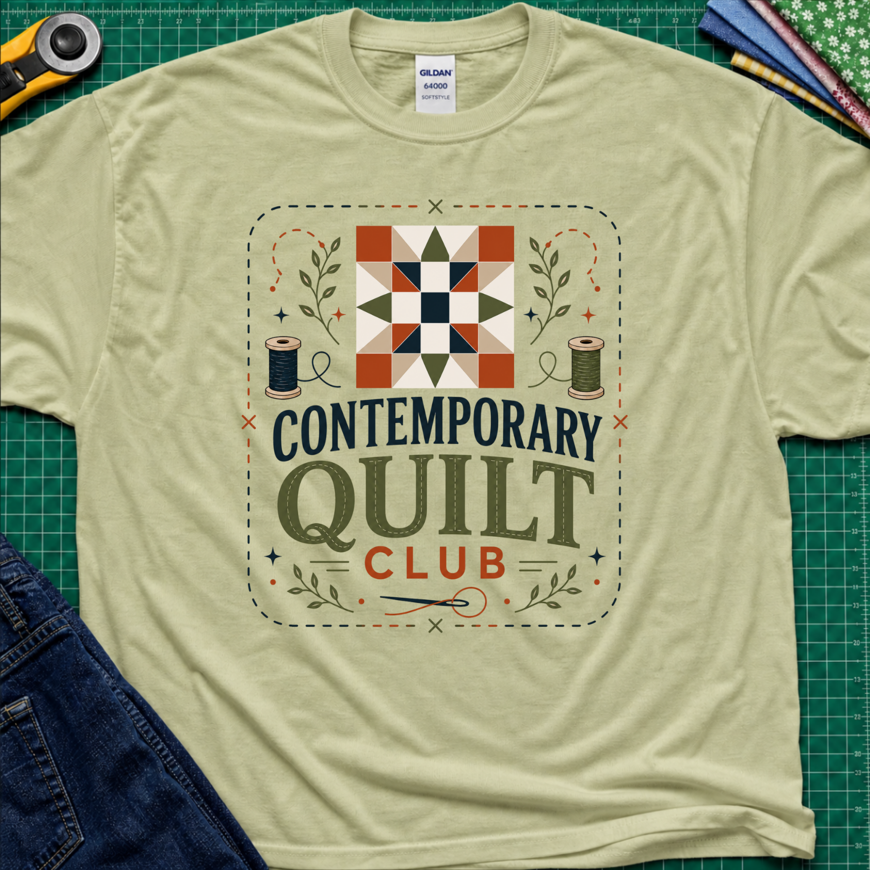 A light pistachio green Gildan t-shirt laid flat on a green cutting mat, featuring a retro-style 'Contemporary Quilt Club' graphic with a quilt pattern, spools of thread, and decorative elements.