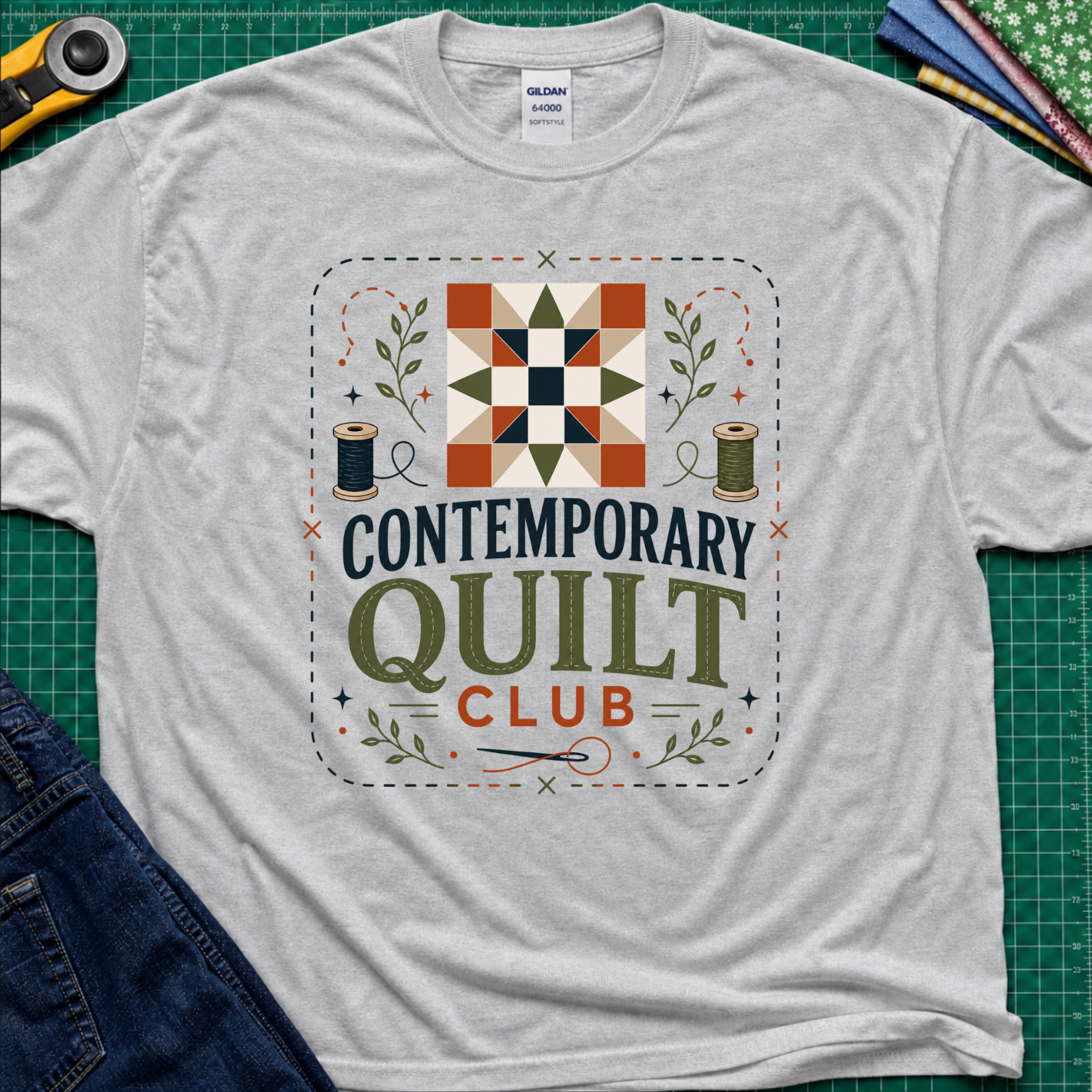 A light grey Gildan t-shirt laid flat on a green cutting mat, featuring a retro-style 'Contemporary Quilt Club' graphic with a quilt pattern, spools of thread, and decorative elements.
