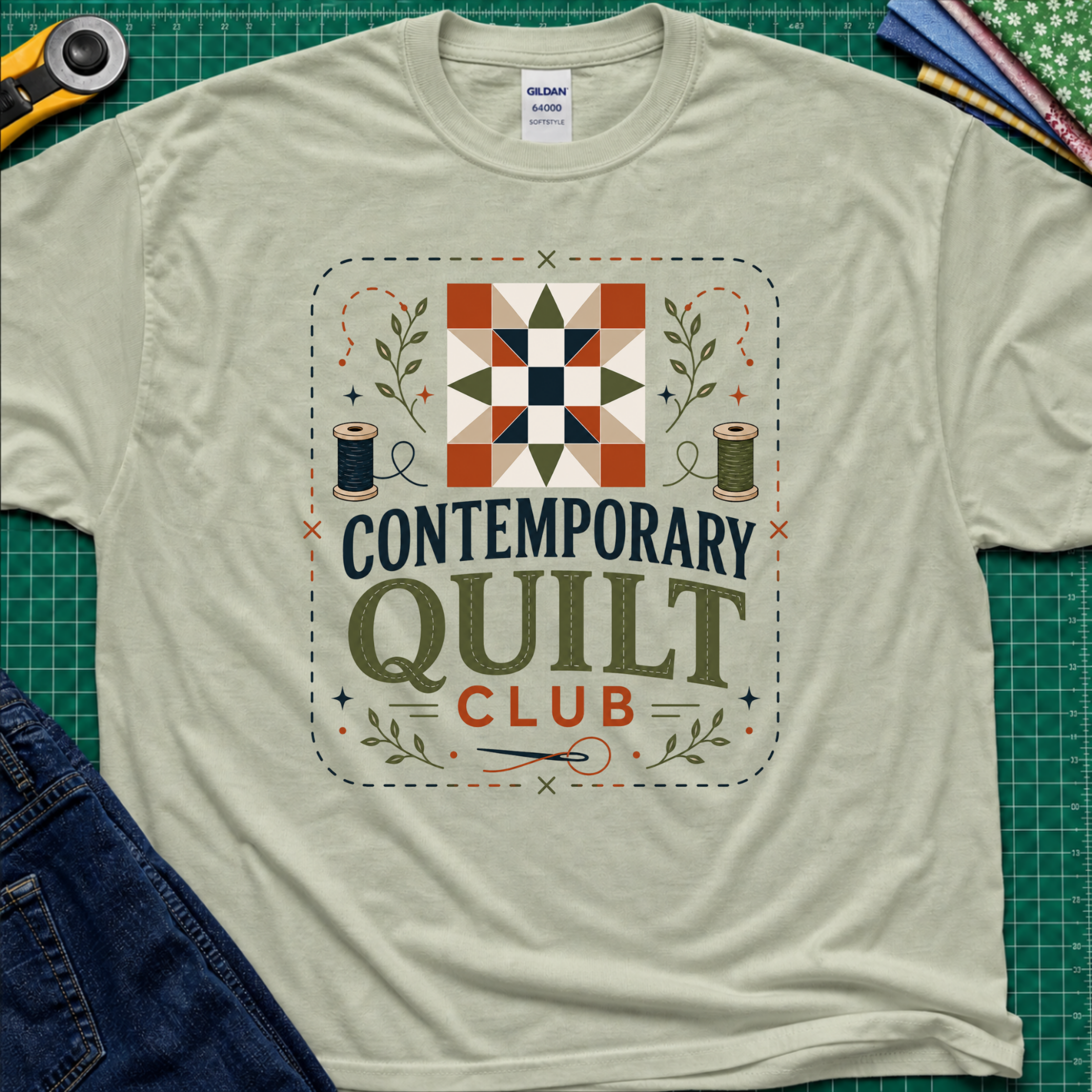 A light sage green Gildan t-shirt laid flat on a green cutting mat, featuring a retro-style 'Contemporary Quilt Club' graphic with a quilt pattern, spools of thread, and decorative elements.