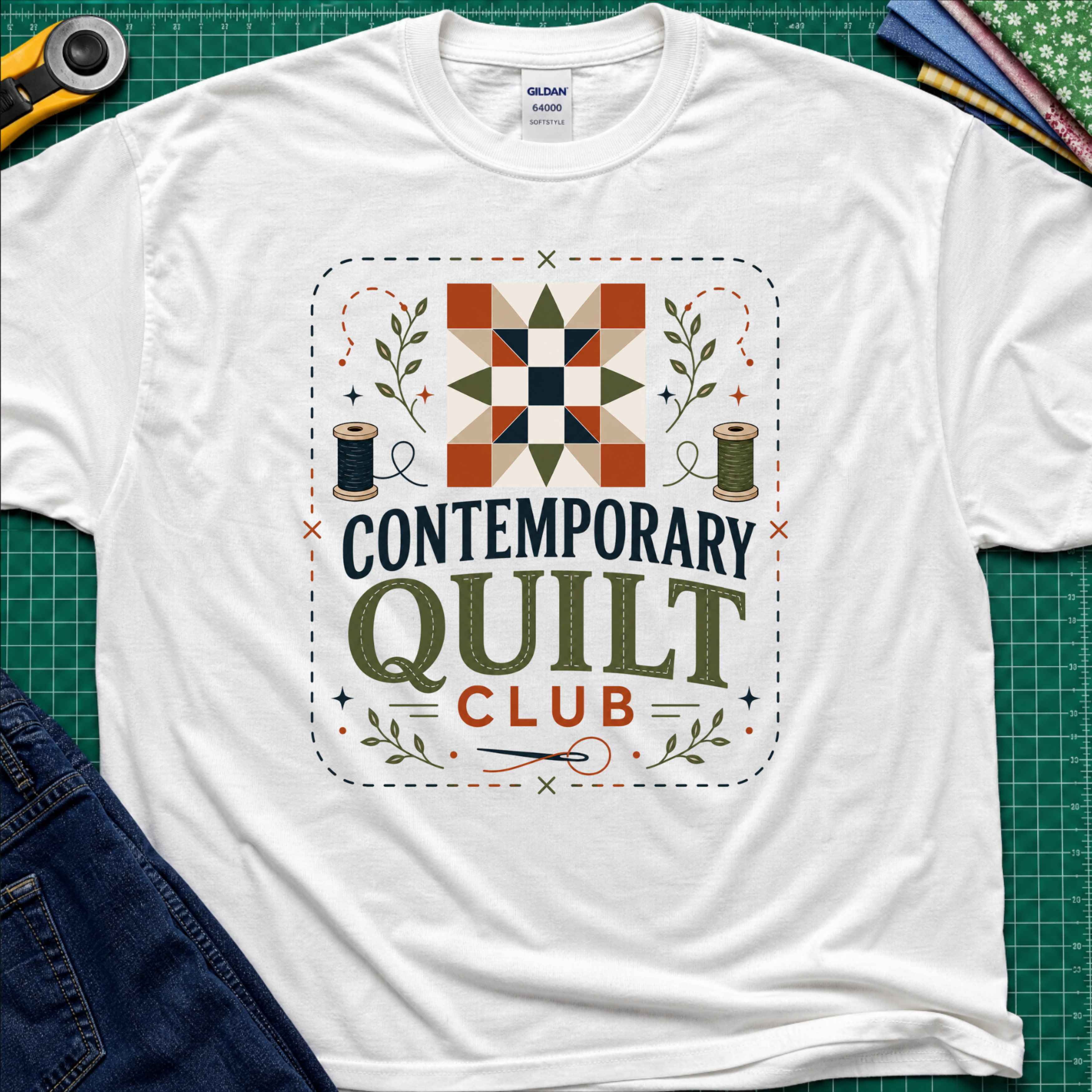 A white Gildan t-shirt laid flat on a green cutting mat, featuring a retro-style 'Contemporary Quilt Club' graphic with a quilt pattern, spools of thread, and decorative elements.