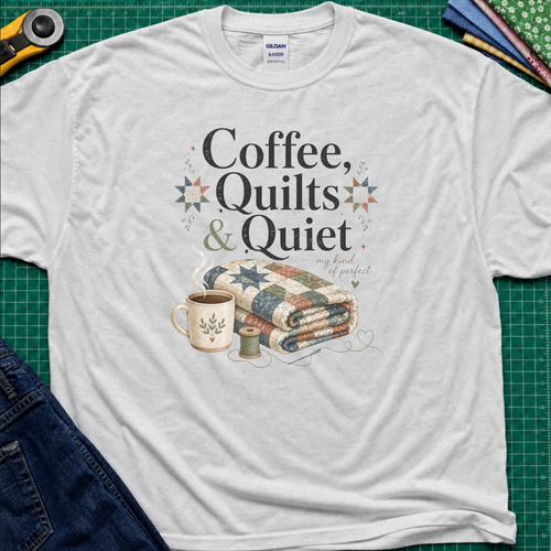 A light grey Gildan t-shirt with the words "Coffee, Quilts and Quiet" above an image of a folded quilt, coffee mug, thread reel and needle. The t-shirt lays flat on a green cutting mat with a rotary cutter, fabric stack and blue jeans.