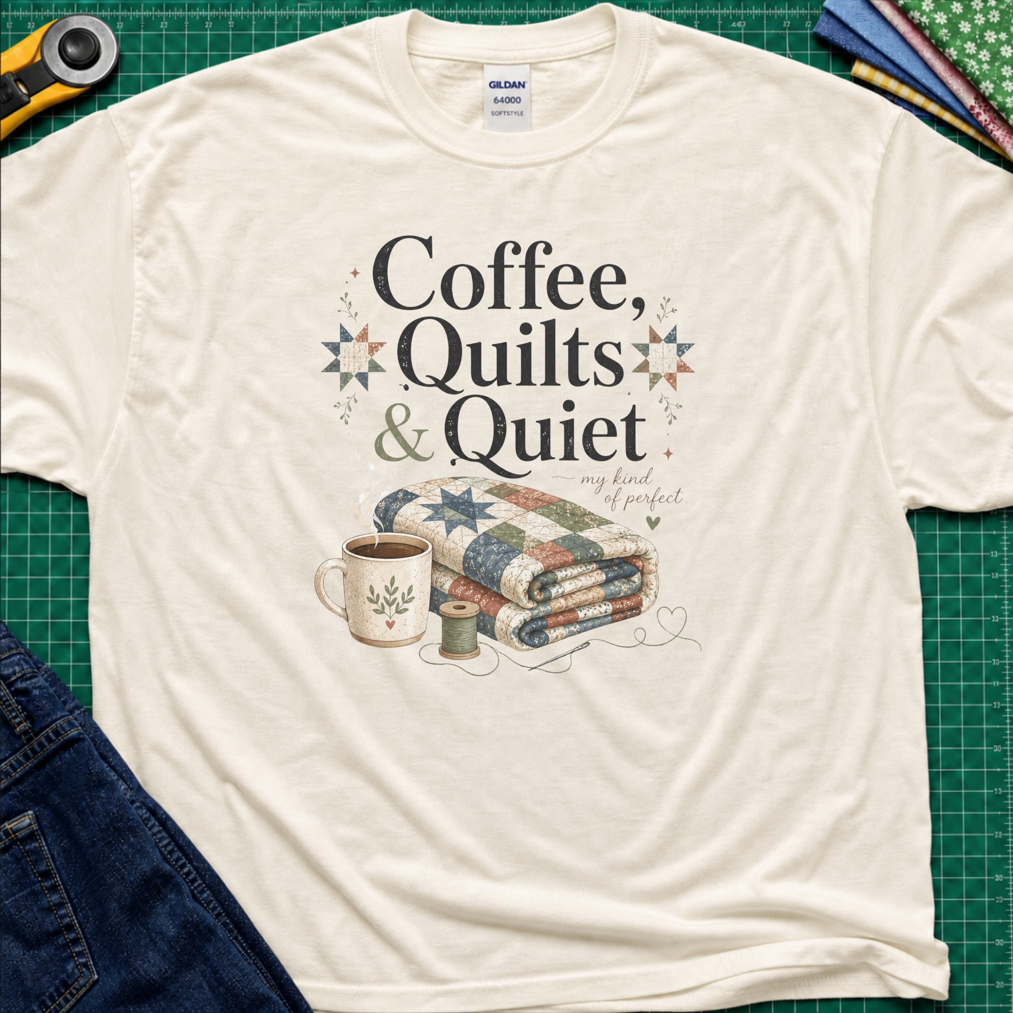 A light cream Gildan t-shirt with the words "Coffee, Quilts and Quiet" above an image of a folded quilt, coffee mug, thread reel and needle. The t-shirt lays flat on a green cutting mat with a rotary cutter, fabric stack and blue jeans.