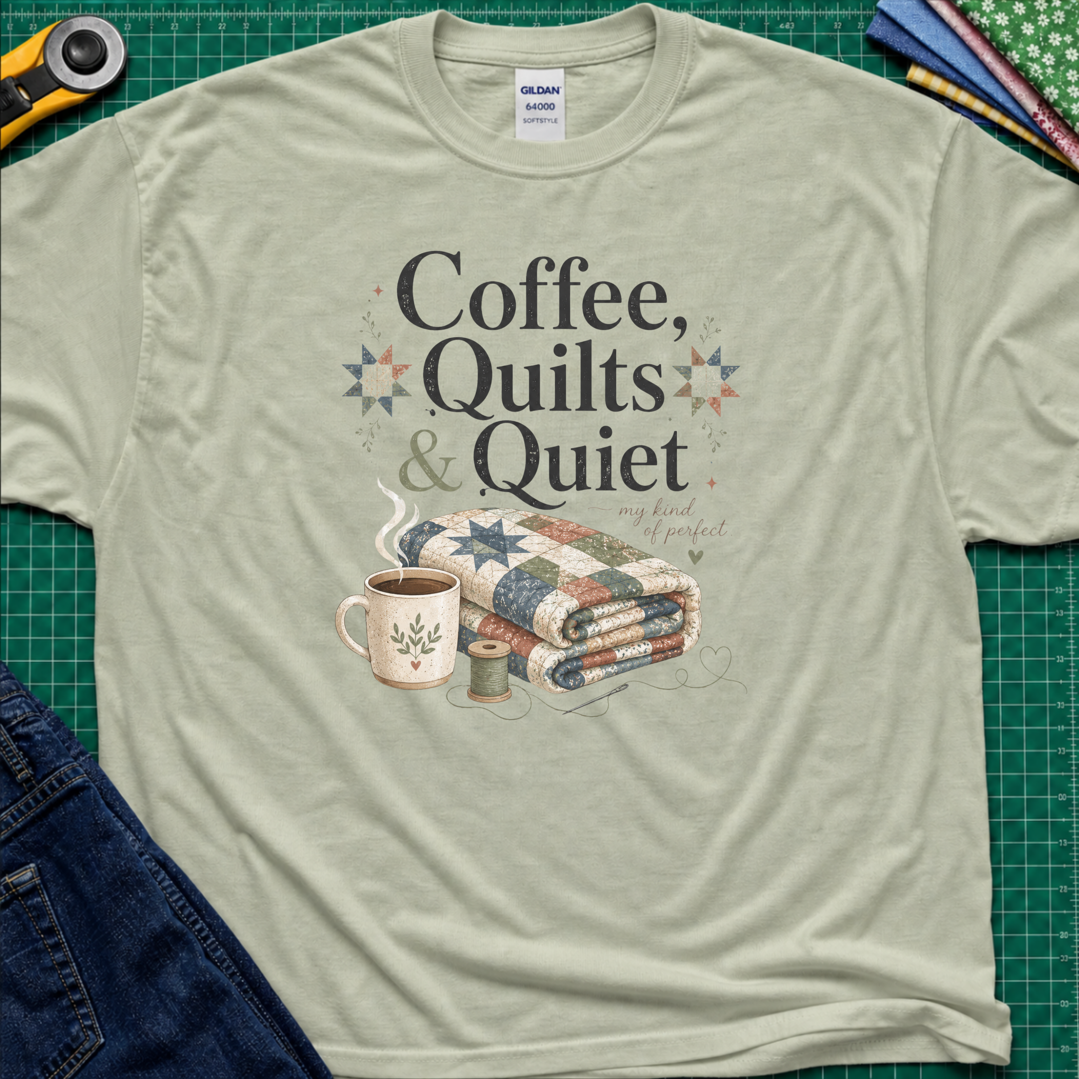 A light sage green Gildan t-shirt with the words "Coffee, Quilts and Quiet" above an image of a folded quilt, coffee mug, thread reel and needle. The t-shirt lays flat on a green cutting mat with a rotary cutter, fabric stack and blue jeans.