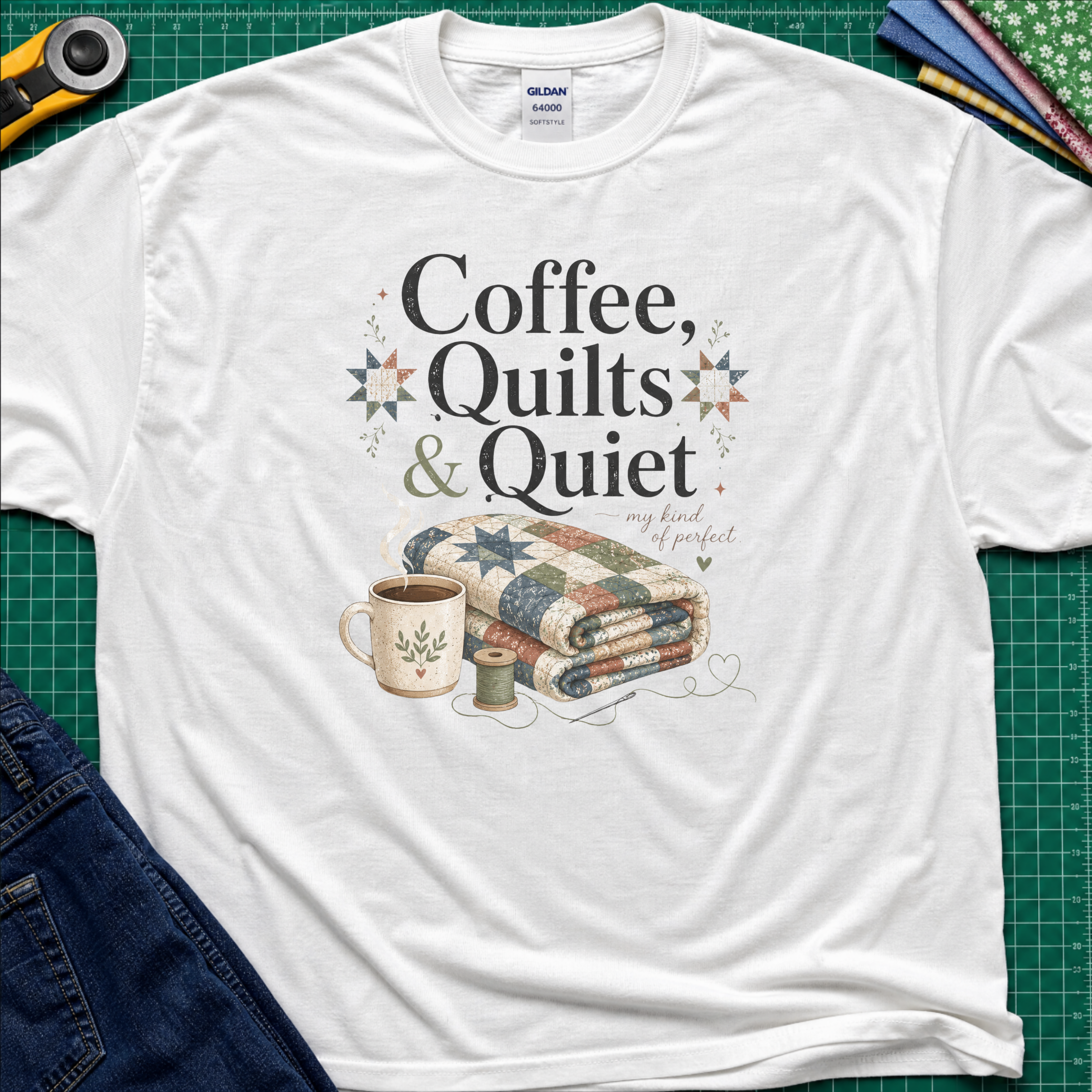 A white Gildan t-shirt with the words "Coffee, Quilts and Quiet" above an image of a folded quilt, coffee mug, thread reel and needle. The t-shirt lays flat on a green cutting mat with a rotary cutter, fabric stack and blue jeans.