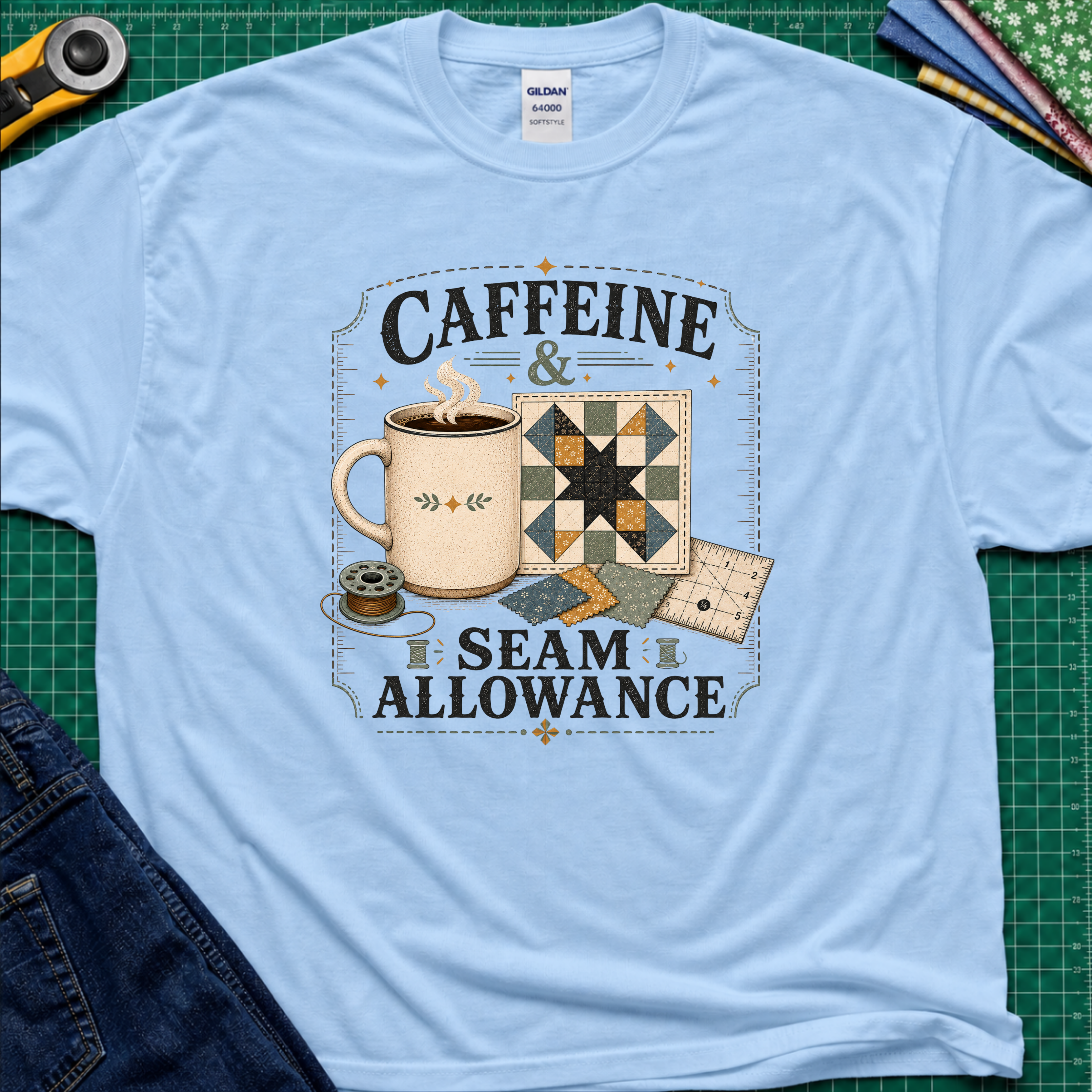 A light blue Gildan t-shirt laid flat on a green cutting mat background, featuring a graphic print that reads 'CAFFEINE & SEAM ALLOWANCE' around an illustration of a coffee mug, sewing thread spool, and quilt blocks.