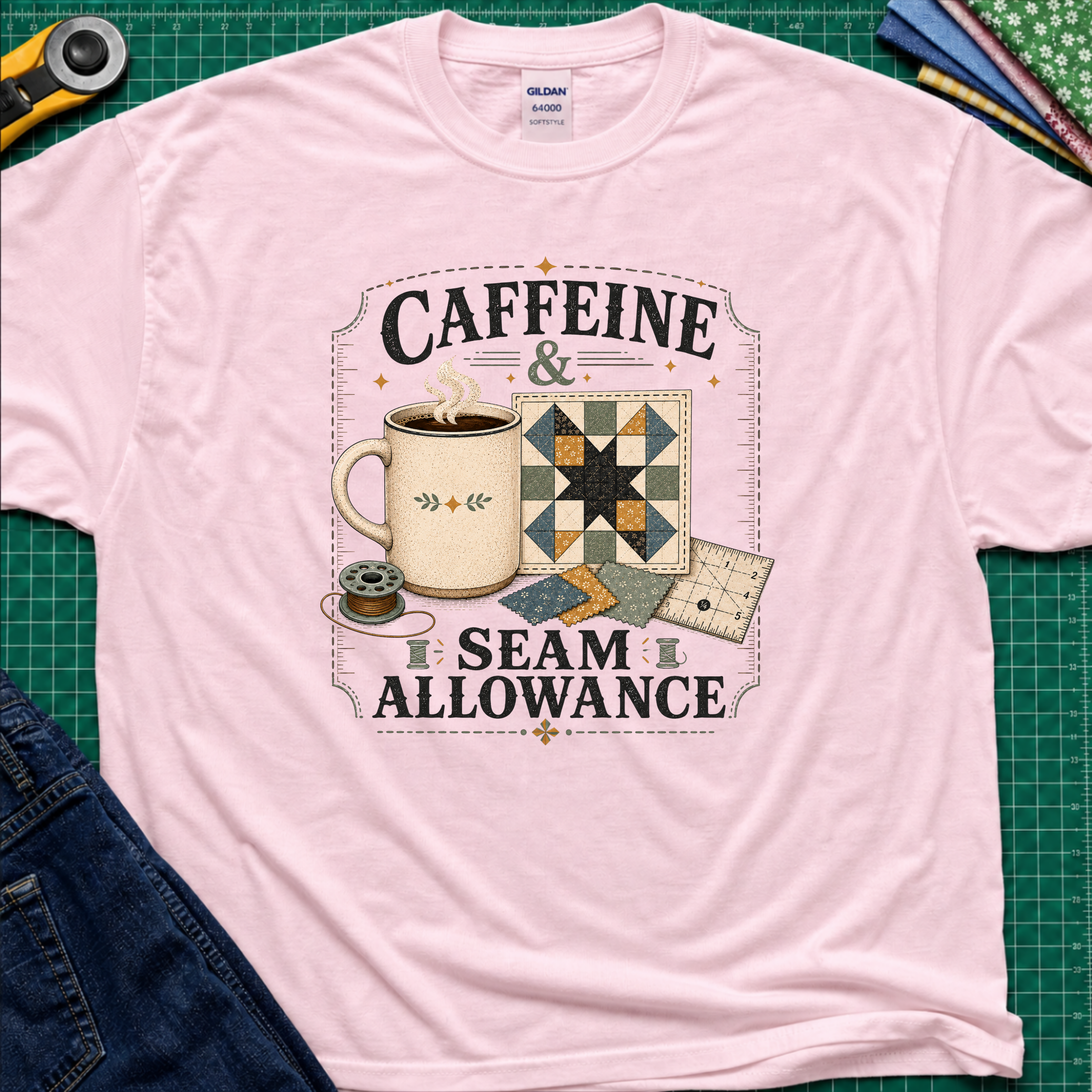 A light pink Gildan t-shirt laid flat on a green cutting mat background, featuring a graphic print that reads 'CAFFEINE & SEAM ALLOWANCE' around an illustration of a coffee mug, sewing thread spool, and quilt blocks.