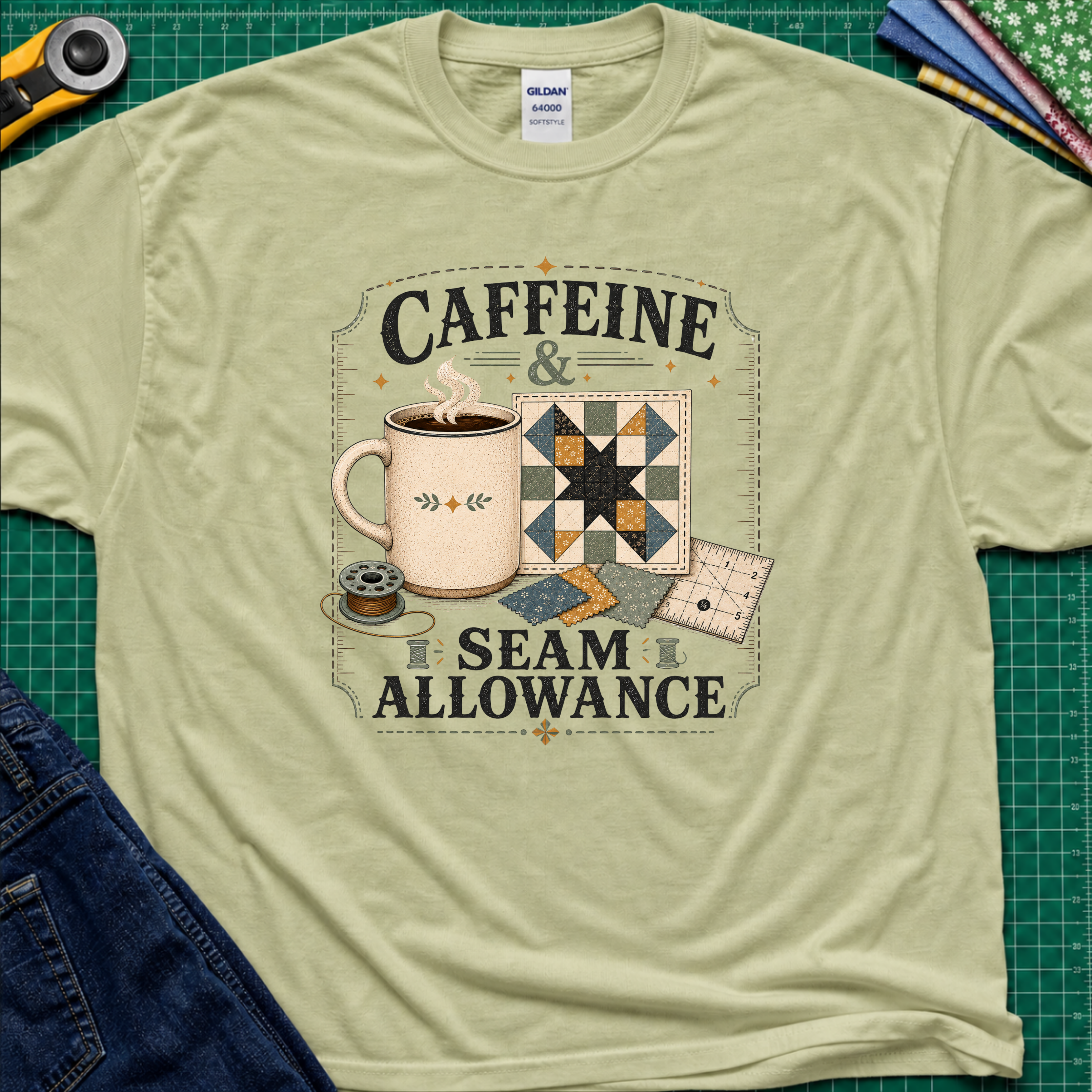A light pistachio green Gildan t-shirt laid flat on a green cutting mat background, featuring a graphic print that reads 'CAFFEINE & SEAM ALLOWANCE' around an illustration of a coffee mug, sewing thread spool, and quilt blocks.