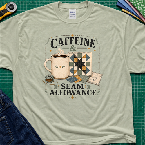 A light sage green Gildan t-shirt laid flat on a green cutting mat background, featuring a graphic print that reads 'CAFFEINE & SEAM ALLOWANCE' around an illustration of a coffee mug, sewing thread spool, and quilt blocks.