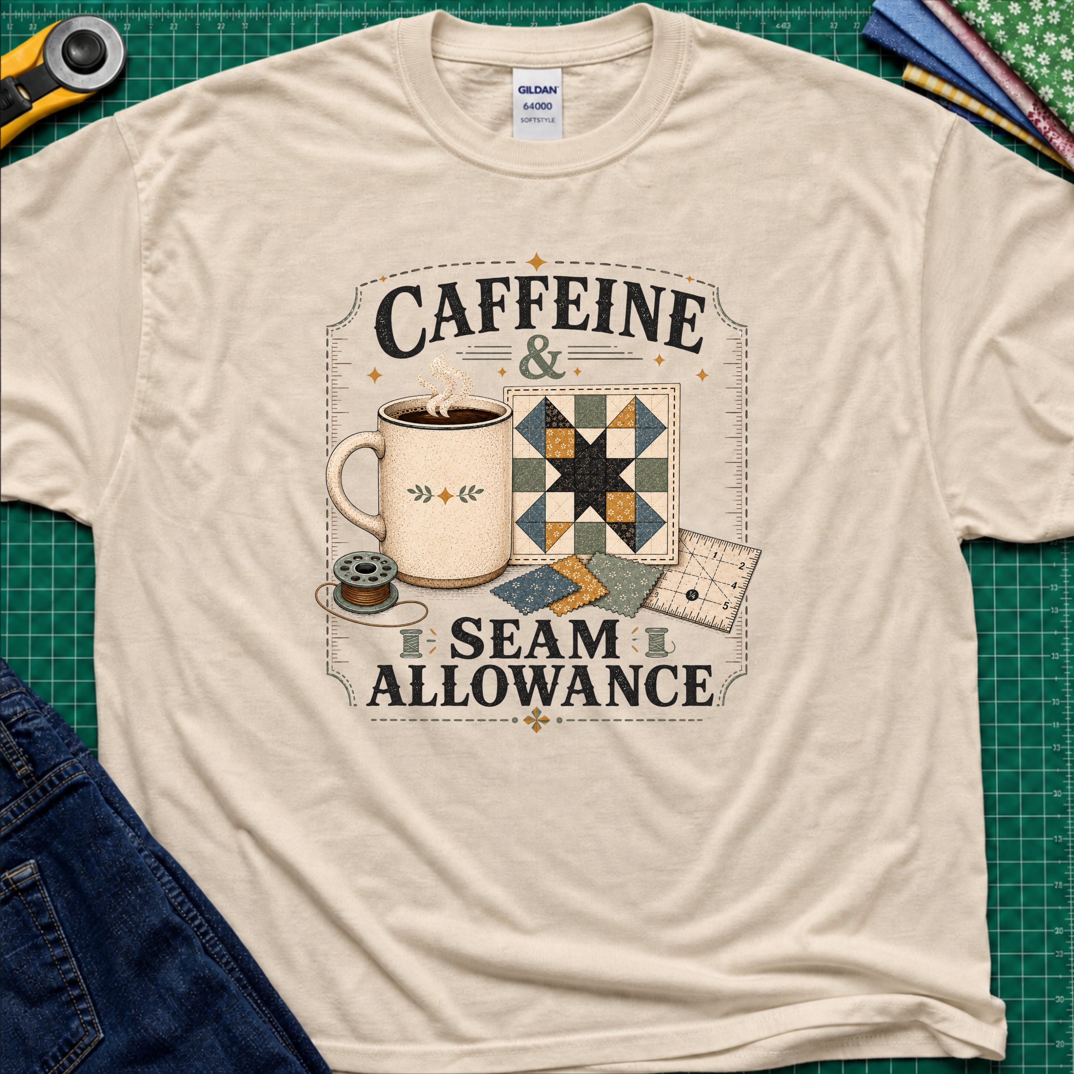 A dark cream Gildan t-shirt laid flat on a green cutting mat background, featuring a graphic print that reads 'CAFFEINE & SEAM ALLOWANCE' around an illustration of a coffee mug, sewing thread spool, and quilt blocks.