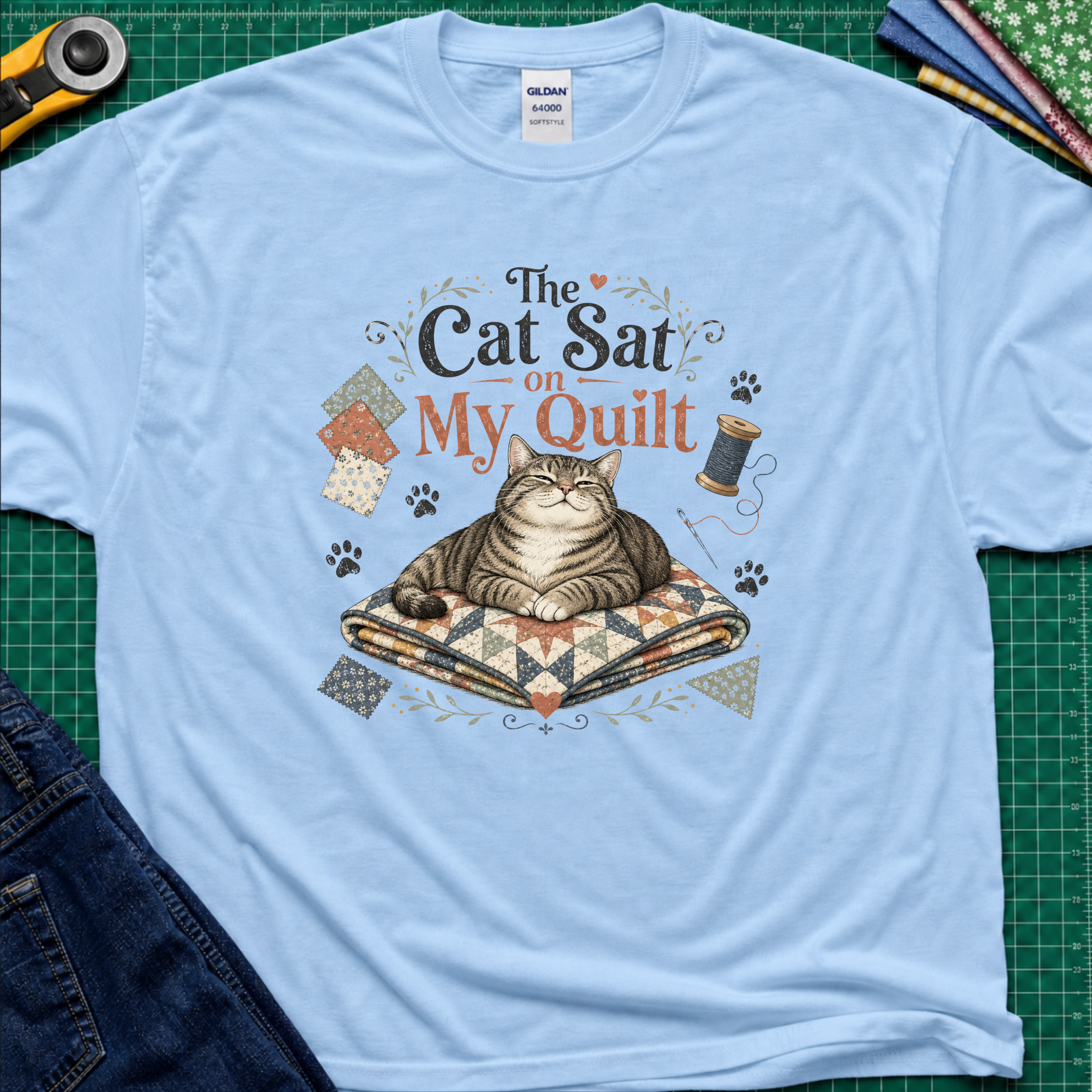 A light blue Gildan t-shirt laid flat on a green cutting mat, featuring a graphic of a tabby cat resting on a quilted blanket with the text 'The Cat Sat on My Quilt' with paw prints and fabric swatches.